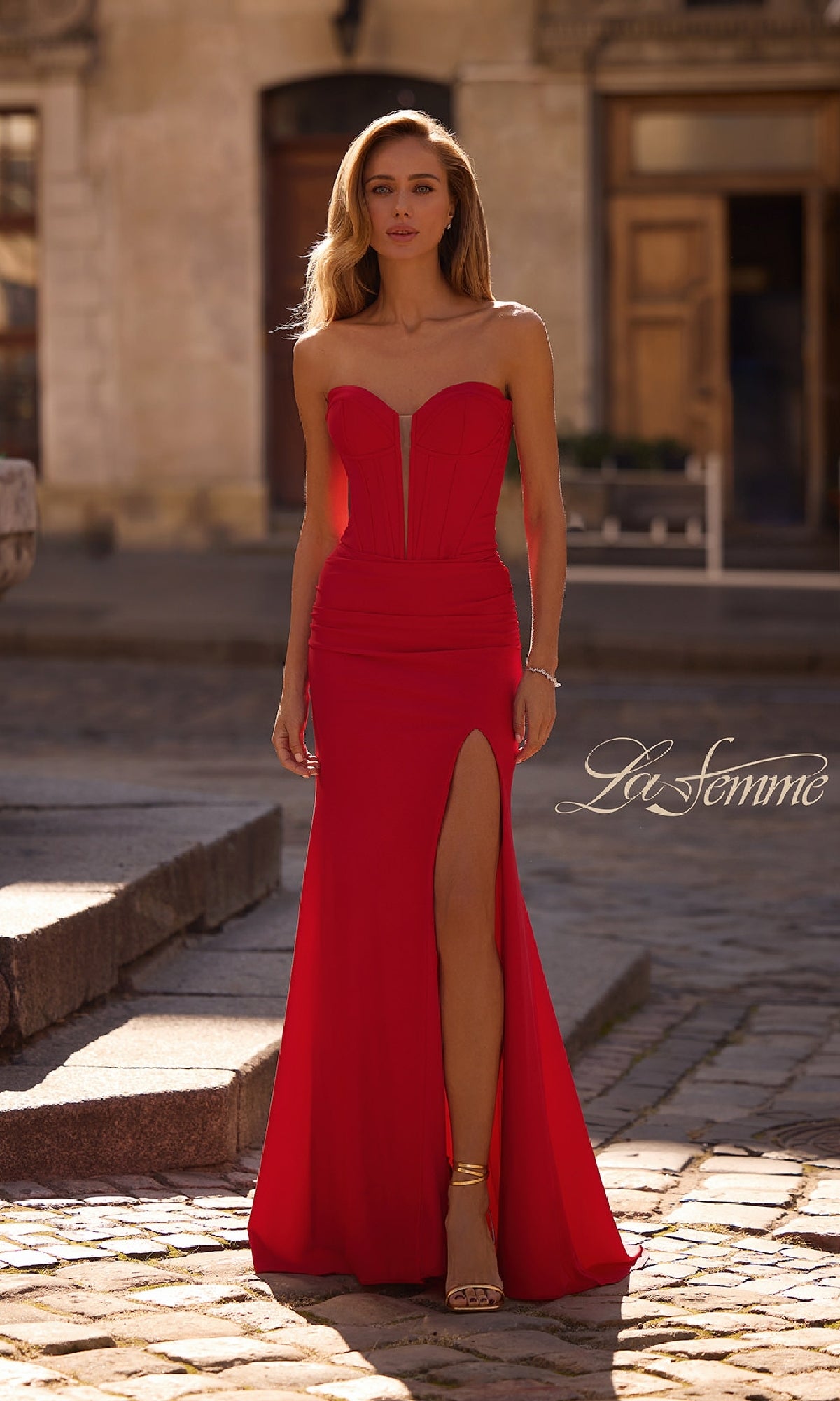 A woman in the La Femme 32941 Strapless Deep V-Neck Prom Gown with a thigh-high slit stands outdoors on a stone path, buildings in the background.