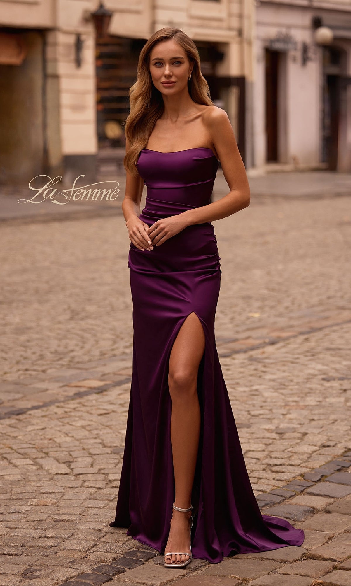 A woman in the Strapless Long Satin Prom Dress: La Femme 32952, featuring a thigh-high slit, stands elegantly on a cobblestone street.