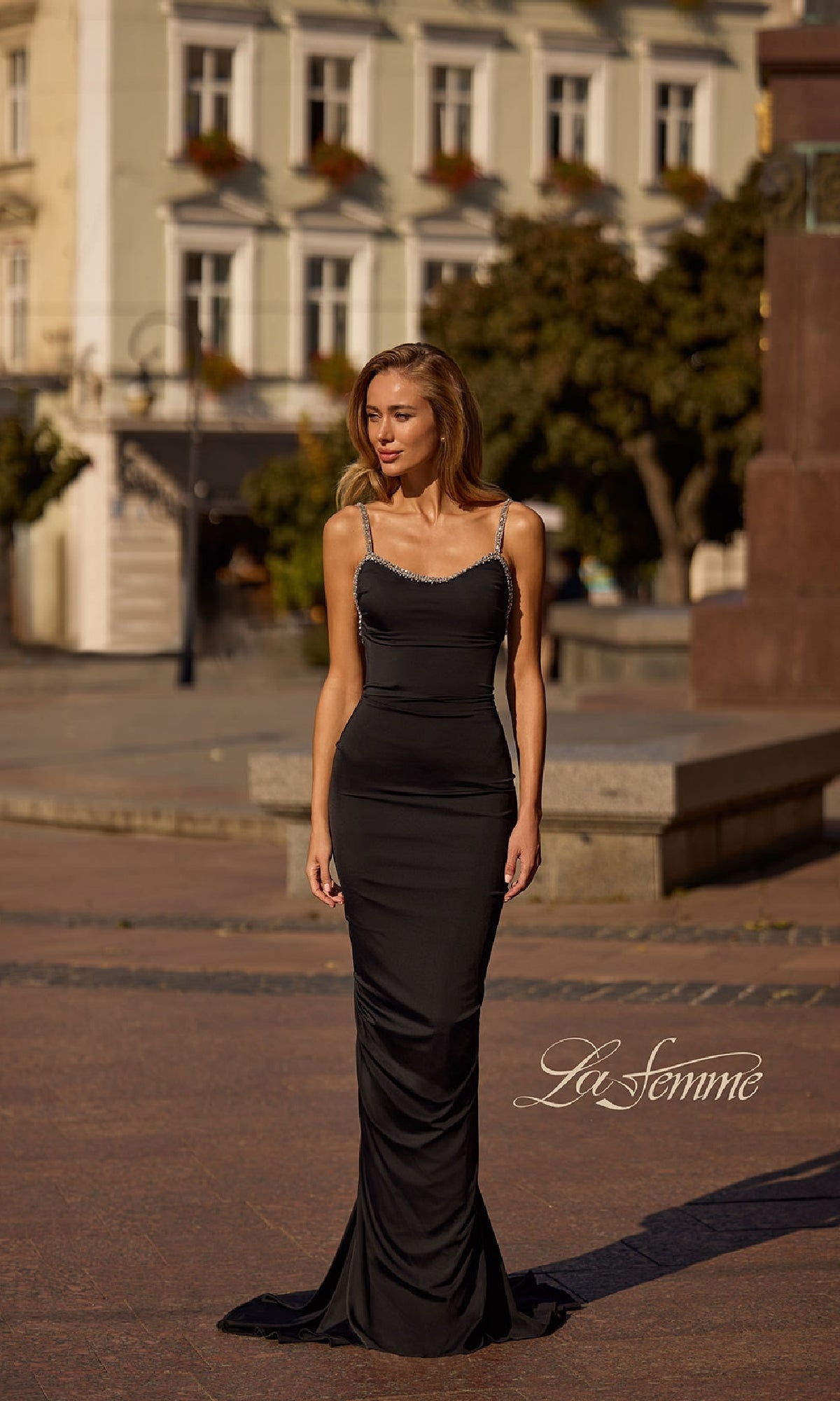 A woman in a fitted black Backless Ruched Long Prom Dress: La Femme 32970 stands outdoors on a paved plaza with buildings and trees in the background. The La Femme logo appears in the lower right corner.