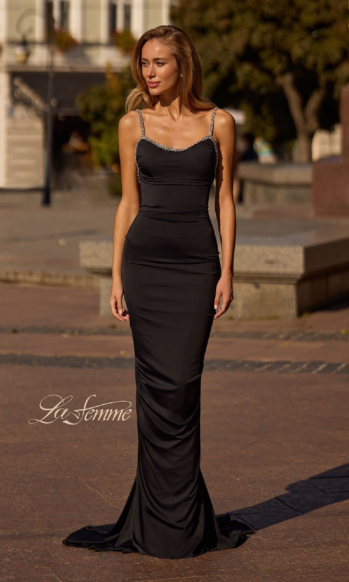 A woman stands outdoors wearing the La Femme 32970 Backless Ruched Long Prom Dress, featuring thin jeweled straps and an elegant fitted black silhouette.
