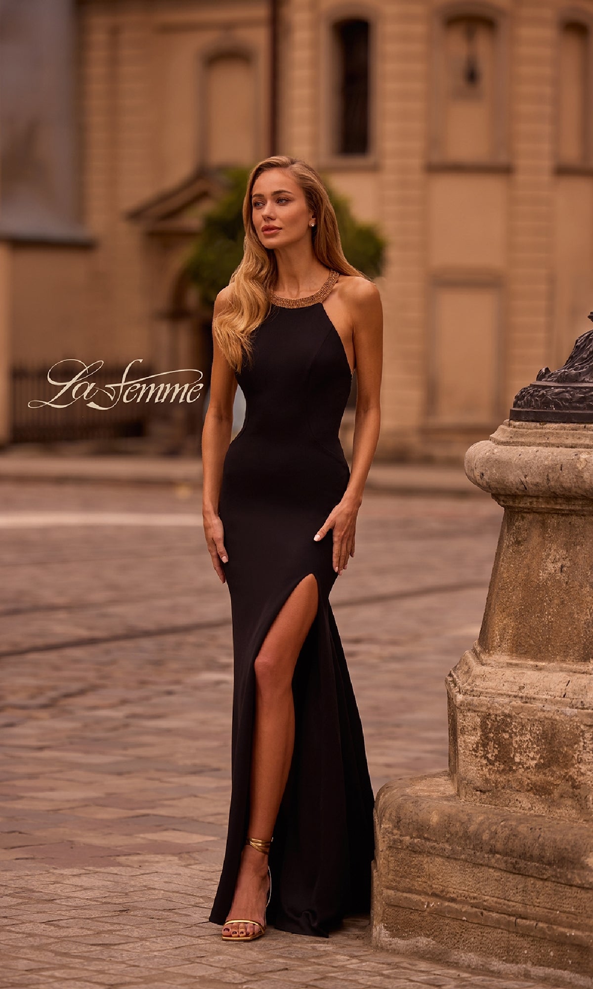A woman stands outdoors on a cobblestone street near a stone structure wearing the La Femme 32976 Gold-Bead Long Black Prom Dress with a thigh-high slit.