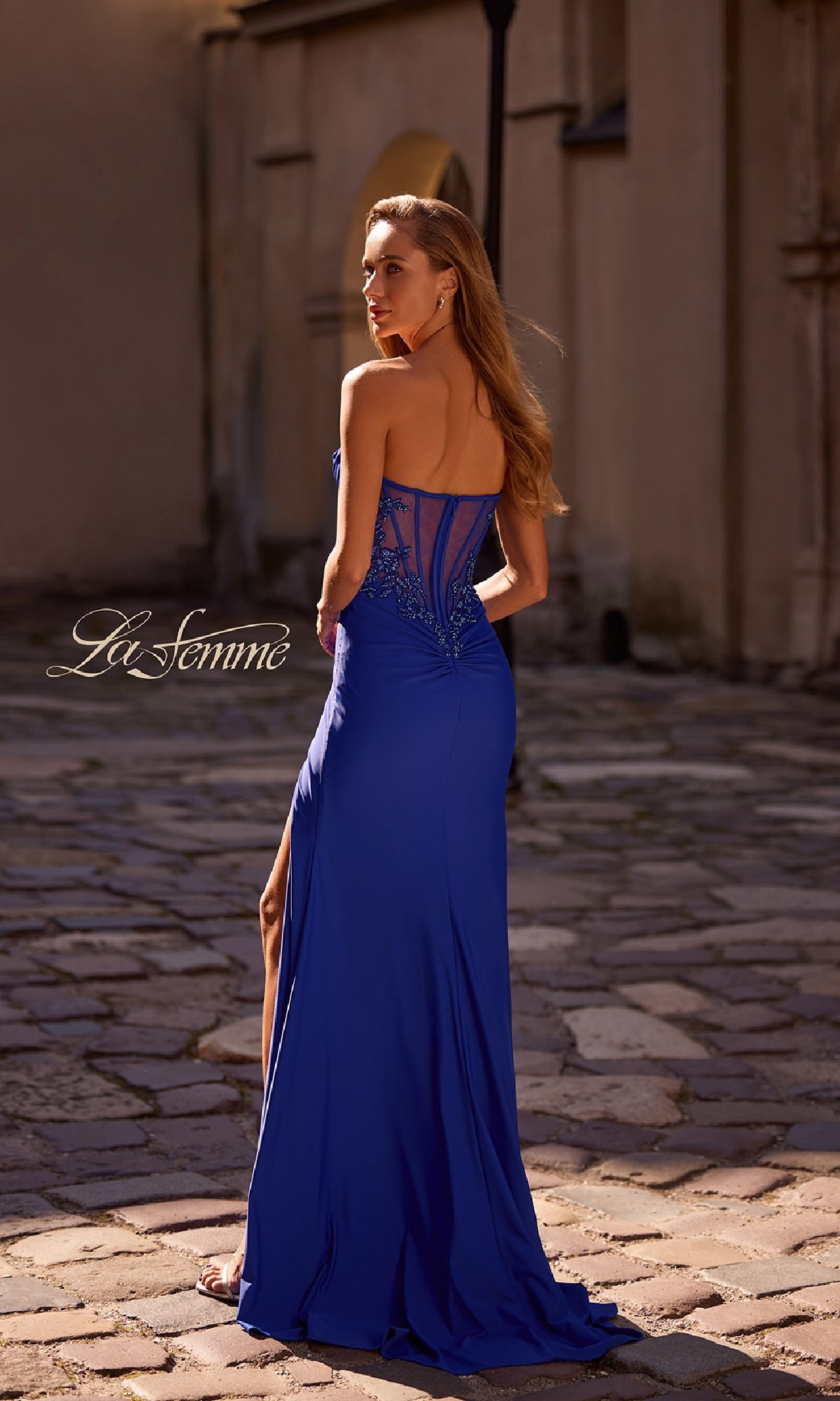 A woman in the La Femme 32981 Beaded-Bodice Long Prom Dress with royal blue lace and beaded details stands on a cobblestone walkway, facing away from the camera.