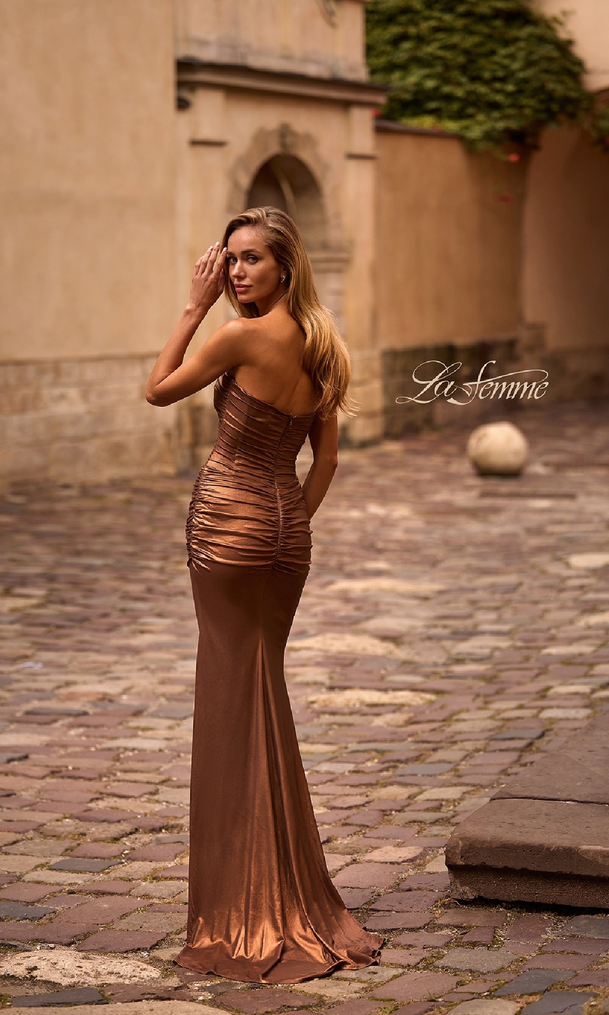 A woman wearing the Metallic Strapless Long Prom Dress: La Femme 33007 stands on a cobblestone path outdoors, glancing over her shoulder.