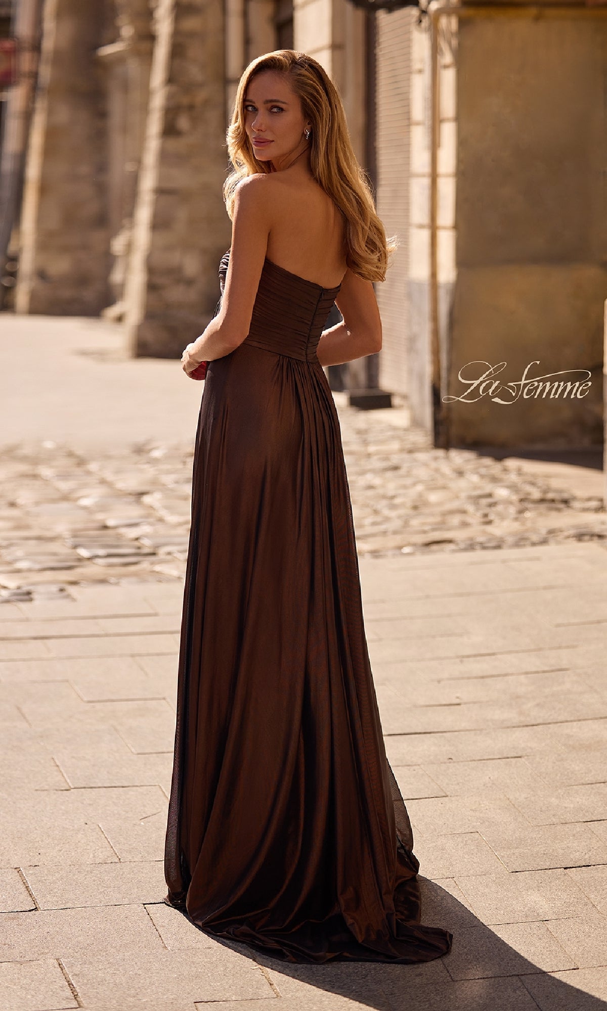 Outdoors on a stone street, a woman looks over her shoulder wearing the La Femme 33047: a strapless, floor-length metallic prom dress.