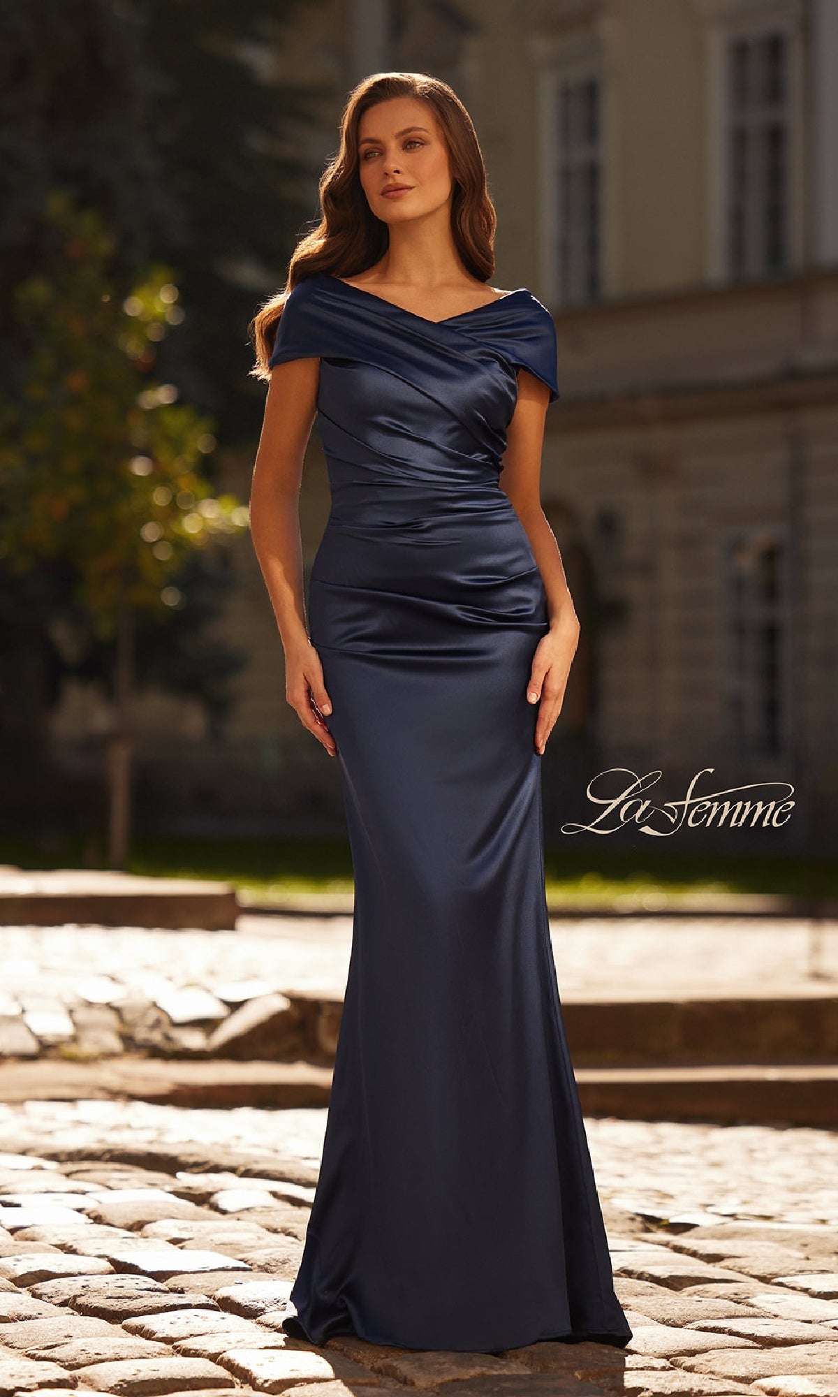 A woman in the Cap-Sleeve Navy Blue Evening Gown: La Femme 33128 stands outdoors on a cobblestone path, with a building and greenery in the background.