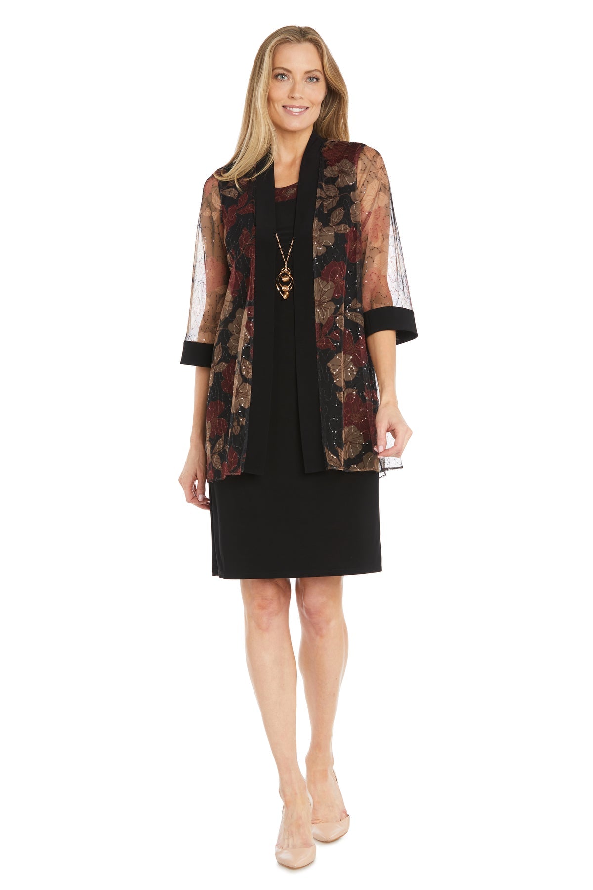 Woman wearing the R&M Richards 3312 Petite Two-Piece Party Dress, featuring a black knee-length semi-formal dress with a sheer floral-patterned jacket, and nude heels, standing and smiling at the camera.