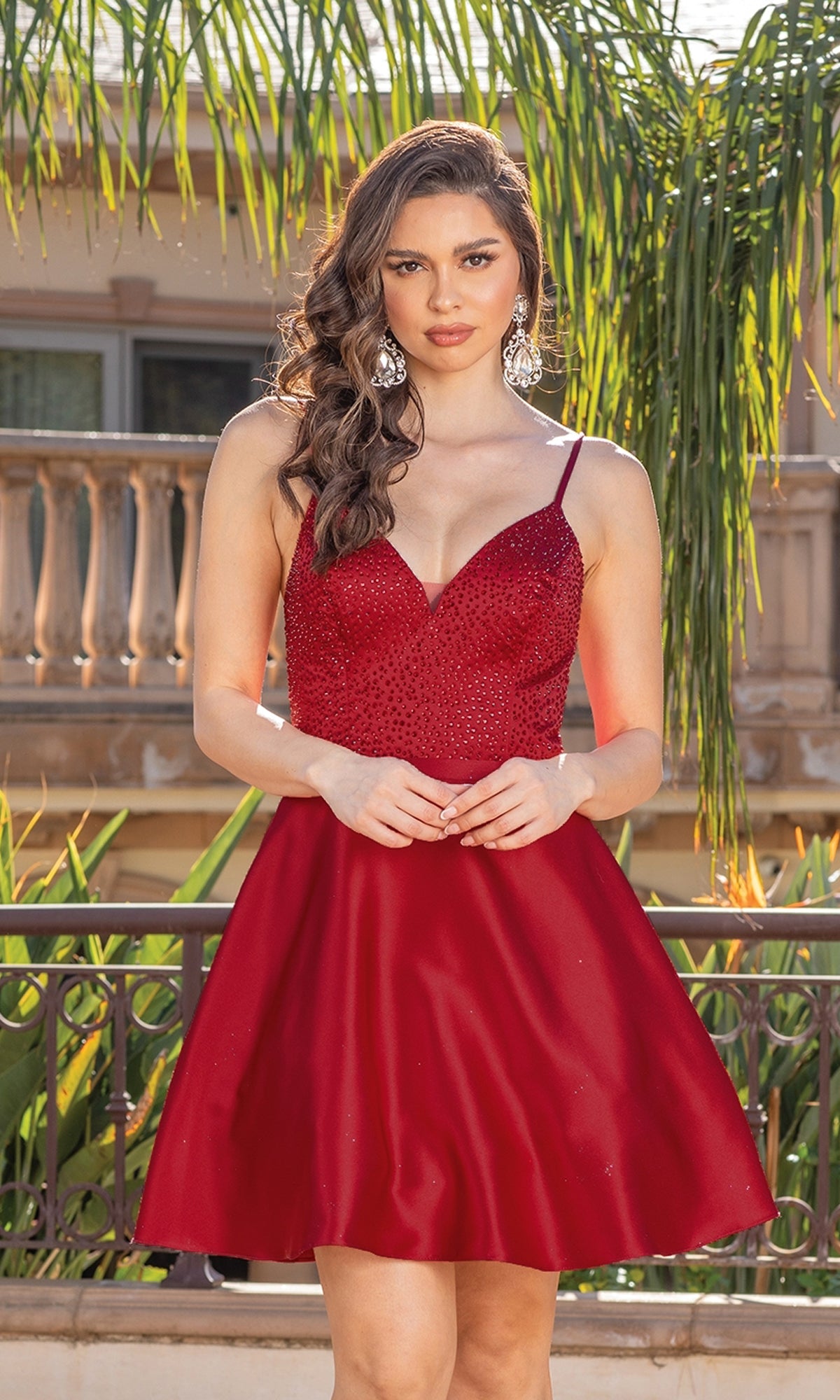 A woman stands outdoors in a sleeveless red Short A-Line Homecoming Dress With Lace-Up Back, featuring a fitted bodice and flared skirt, styled with large earrings. Green foliage and a building appear in the background.