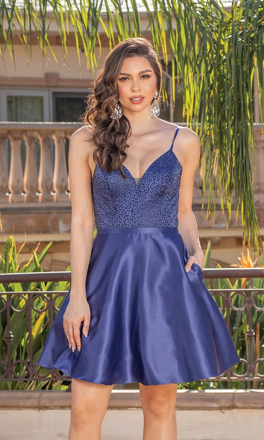 A woman poses outside in a Short A-Line Homecoming Dress With Lace-Up Back, featuring a deep blue satin finish, beaded v-neck bodice, and flared skirt, set against greenery and a balcony railing.