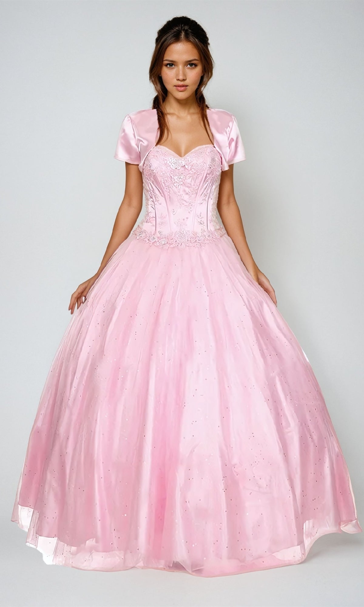 A young woman poses against a plain background in the Juliet 332 Long Prom Dress, a light pink, floor-length ball gown featuring a sweetheart neckline, short sleeves, and a fitted embellished bodice.