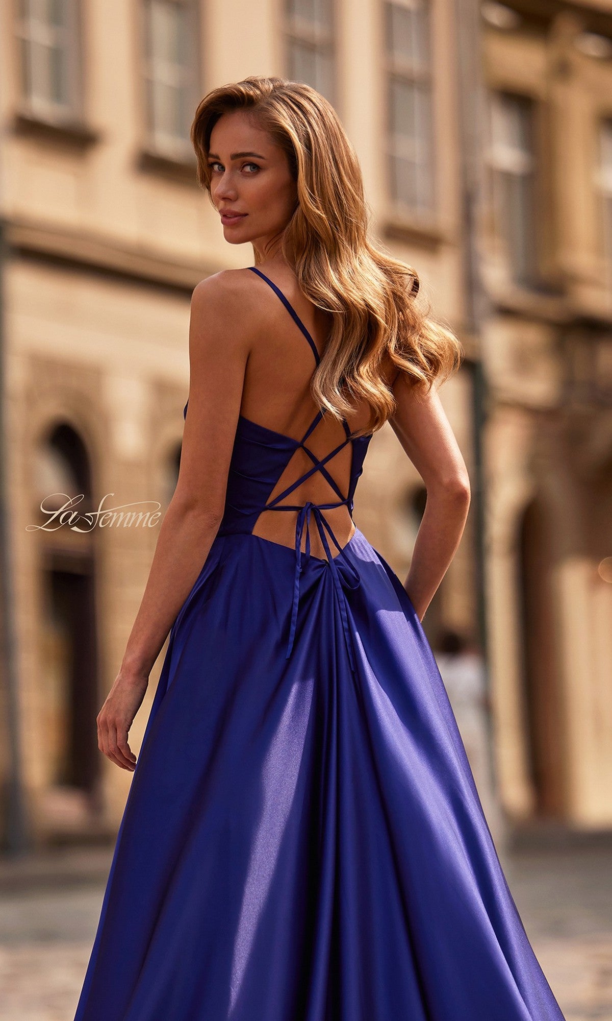 A woman wearing the La Femme 33373 long prom dress in royal blue with an a-line silhouette, high leg slit, and open strappy back stands on a city street, looking over her shoulder.