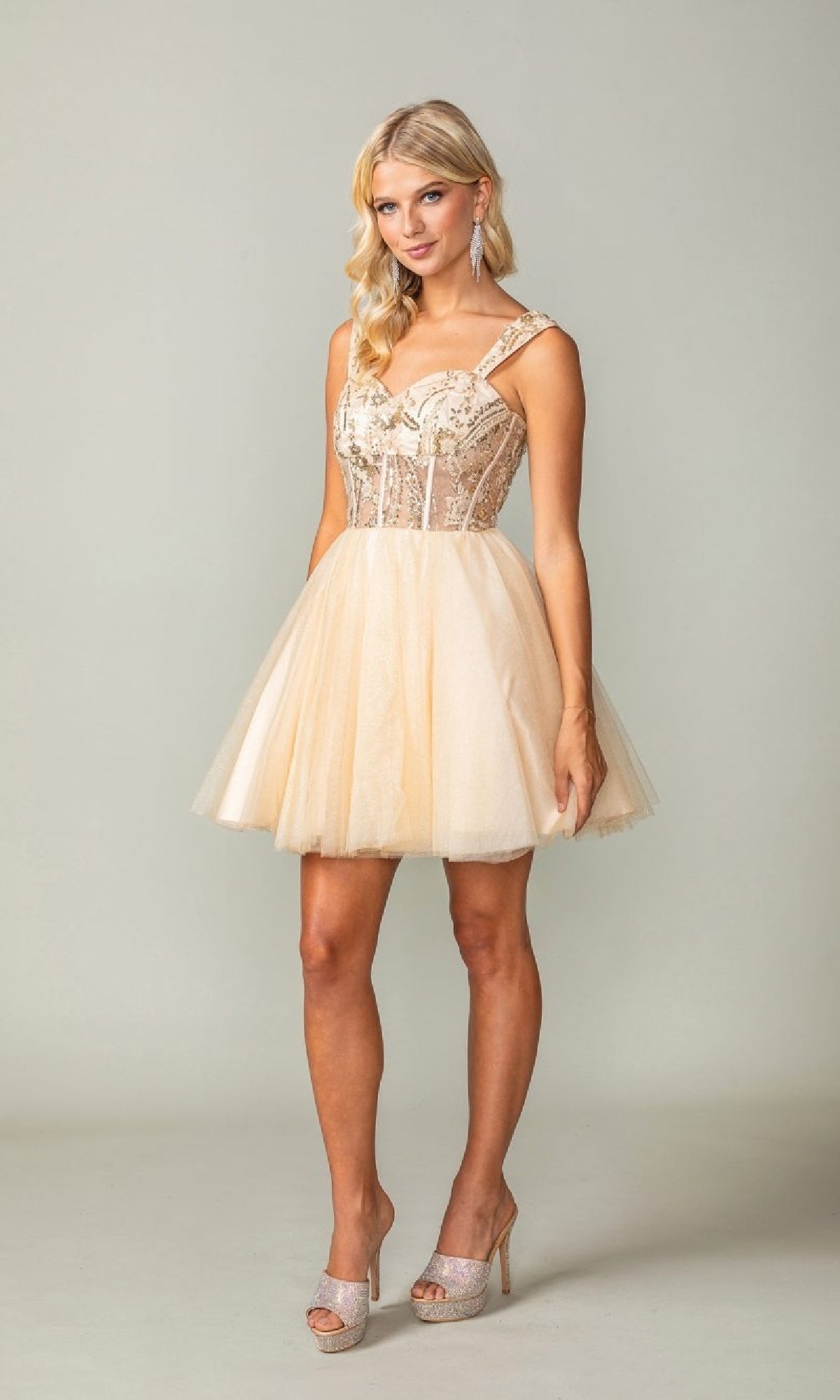 A woman wears the Lace-Up Open-Back Hoco Dress: Dancing Queen 3375, a short light beige tulle homecoming dress with lace details and one-shoulder straps, paired with sparkling heels against a plain background.