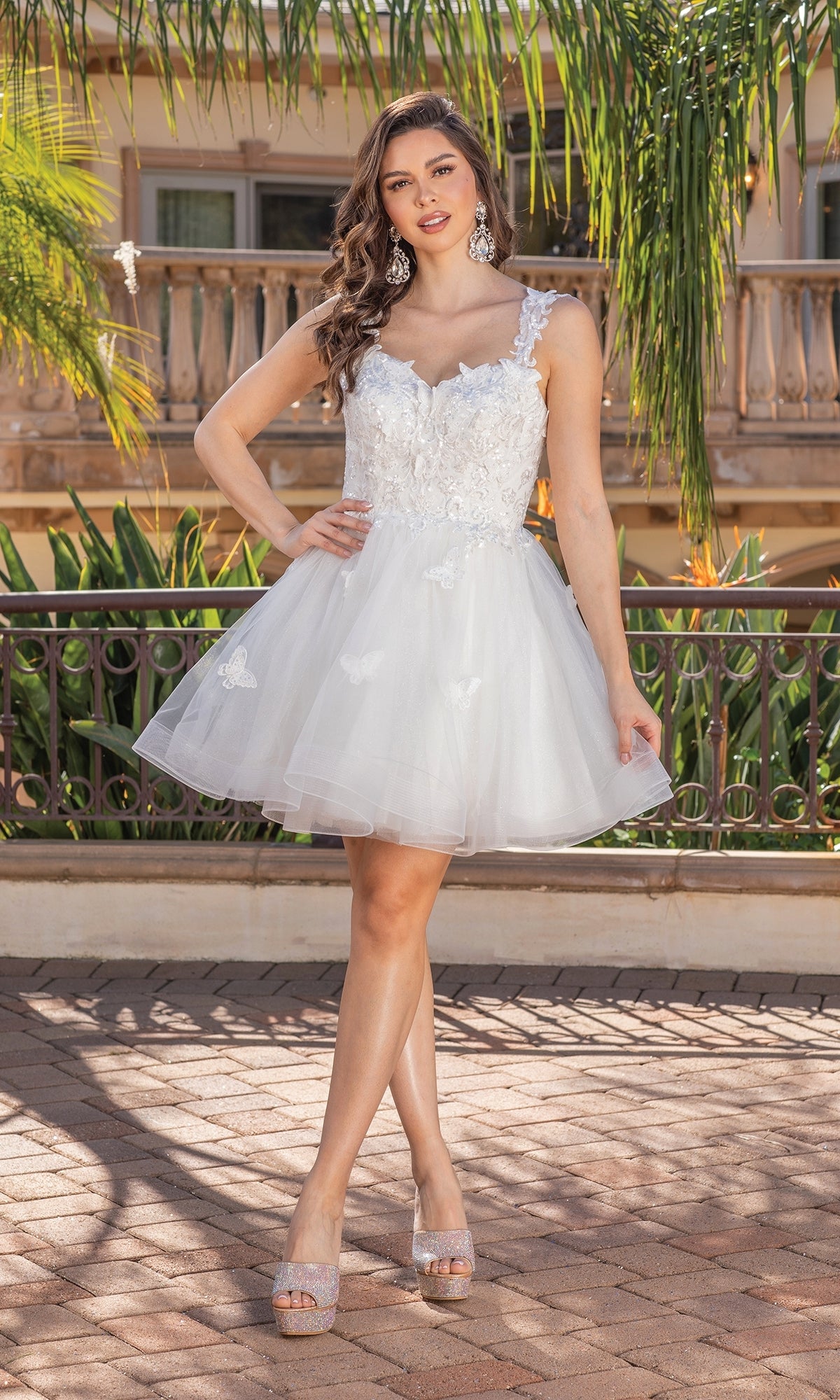 A woman stands outdoors in the Embroidered-Lace Short Babydoll Hoco Dress 3338, featuring white lace, tulle, and floral accents. She completes her look with large earrings and platform heels amid palm trees and a stone railing.