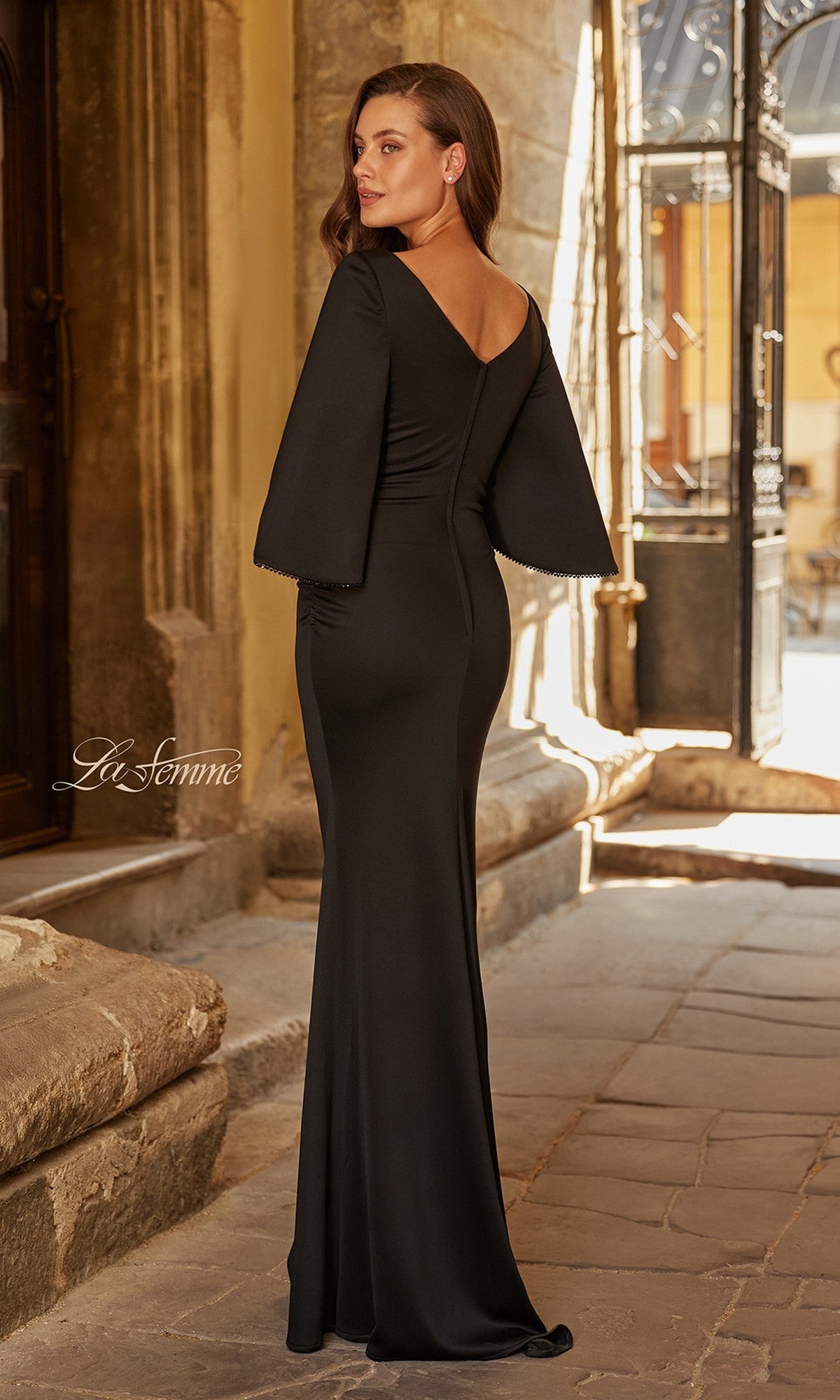 A woman wearing the La Femme 33391 long formal dress with cape-style sleeves and beaded trim stands on a stone path in front of an old building, facing away from the camera.