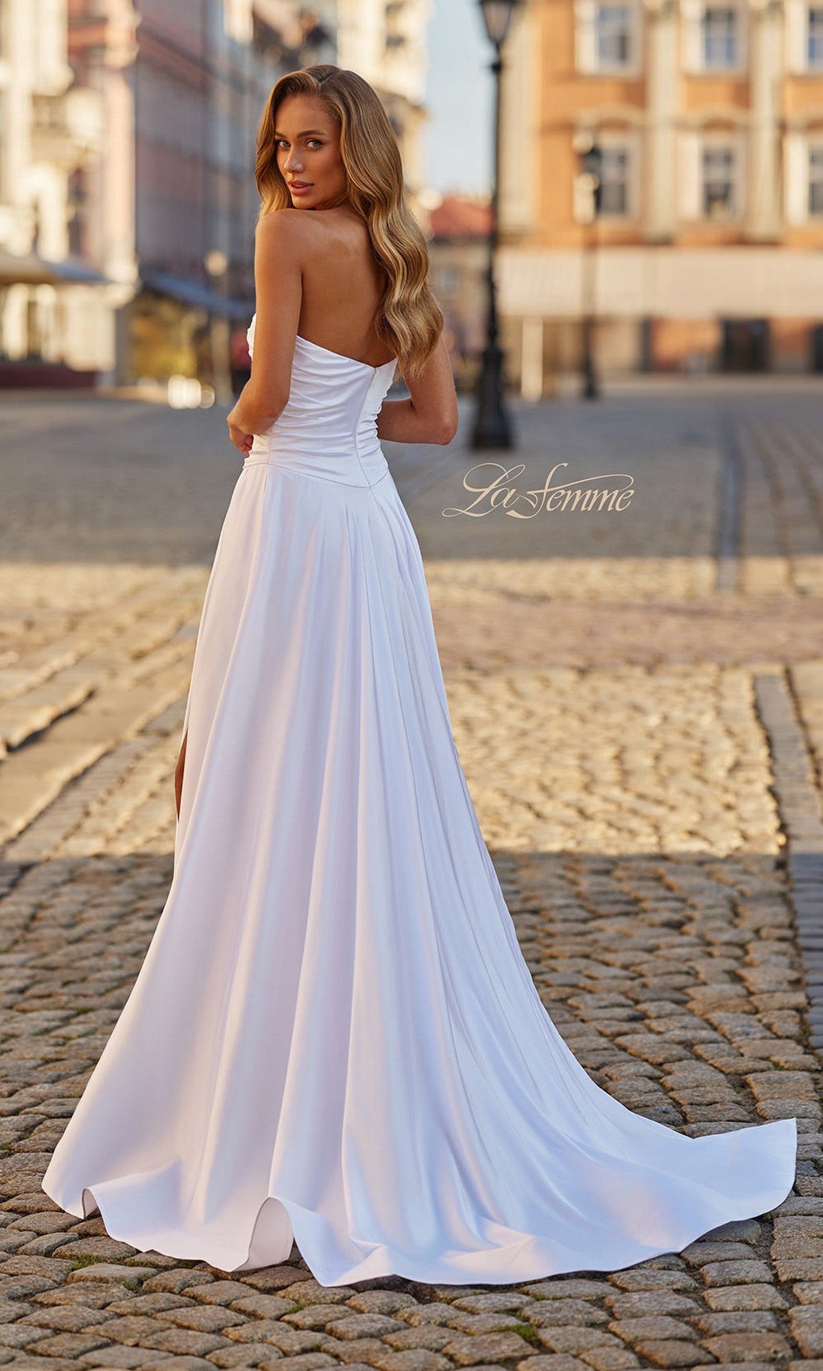 A woman wearing the Strapless Sweetheart Prom Gown: La Femme 33417 stands on a cobblestone street, glancing over her shoulder as the elegant white, floor-length dress flows and buildings rise behind her.