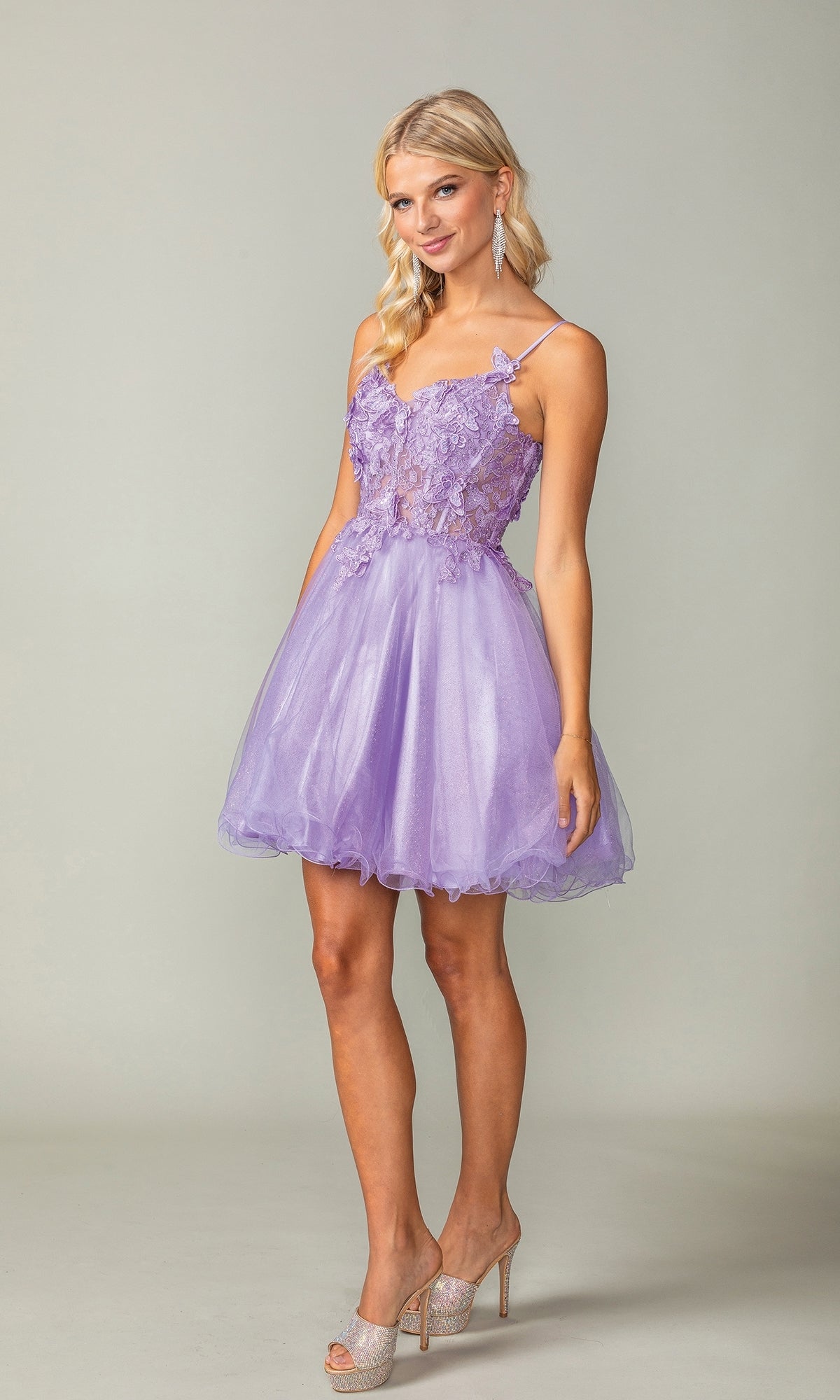 A woman poses in the Lace-Up Short Butterfly Homecoming Dress 3341—light purple with lace details and a tulle skirt—paired with silver heels, set against a plain background.
