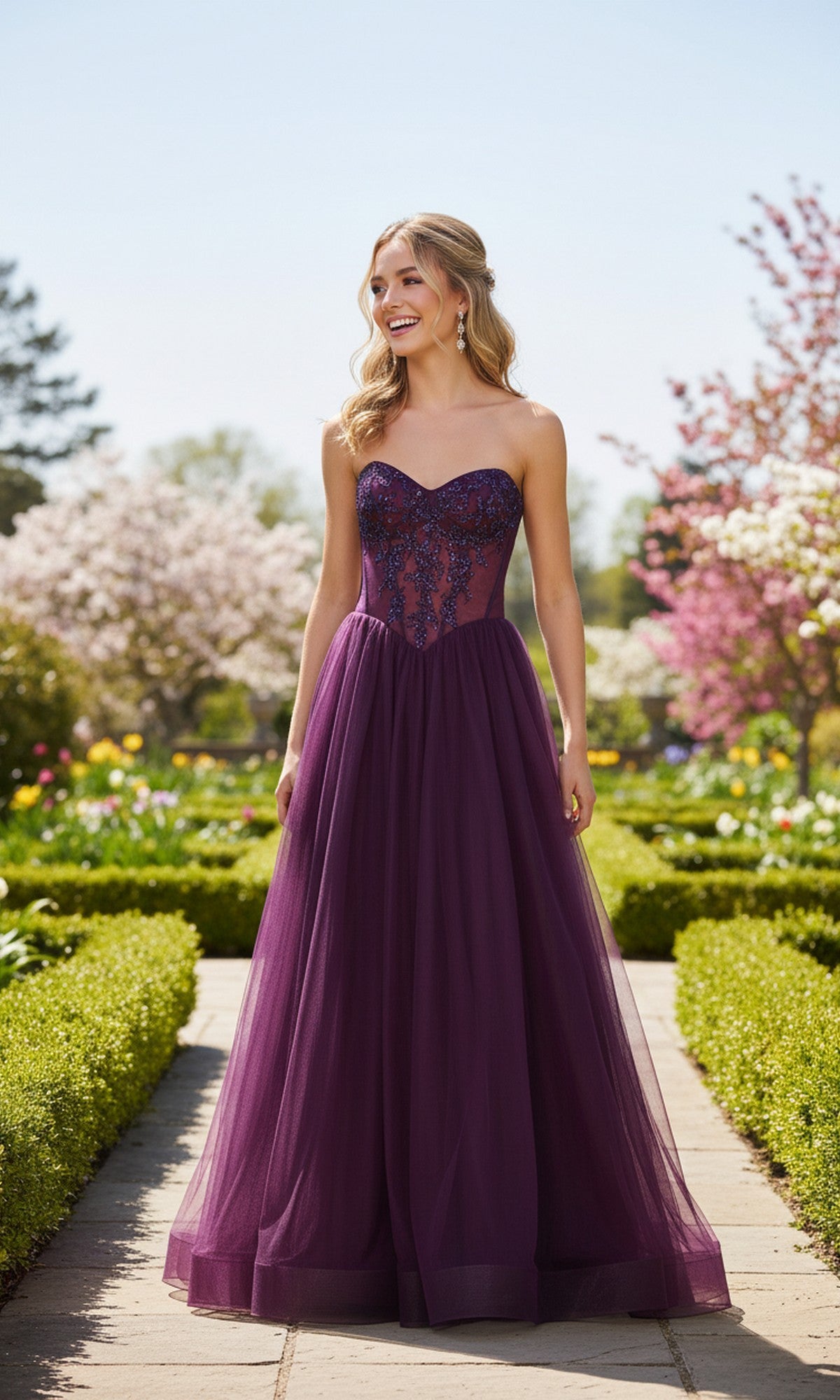 A woman in the Strapless Sheer-Corset Prom Gown: La Femme 33491 stands on a garden pathway, surrounded by blooming trees and lush greenery.