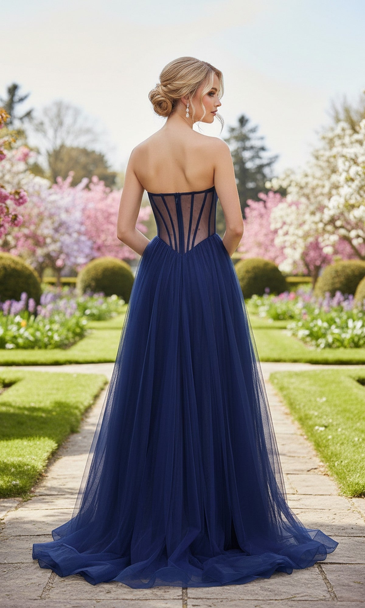 A woman in the Strapless Sheer-Corset Prom Gown: La Femme 33491, with its long A-line silhouette and embroidered corset bodice, stands on a garden path lined with blooming trees and manicured bushes.