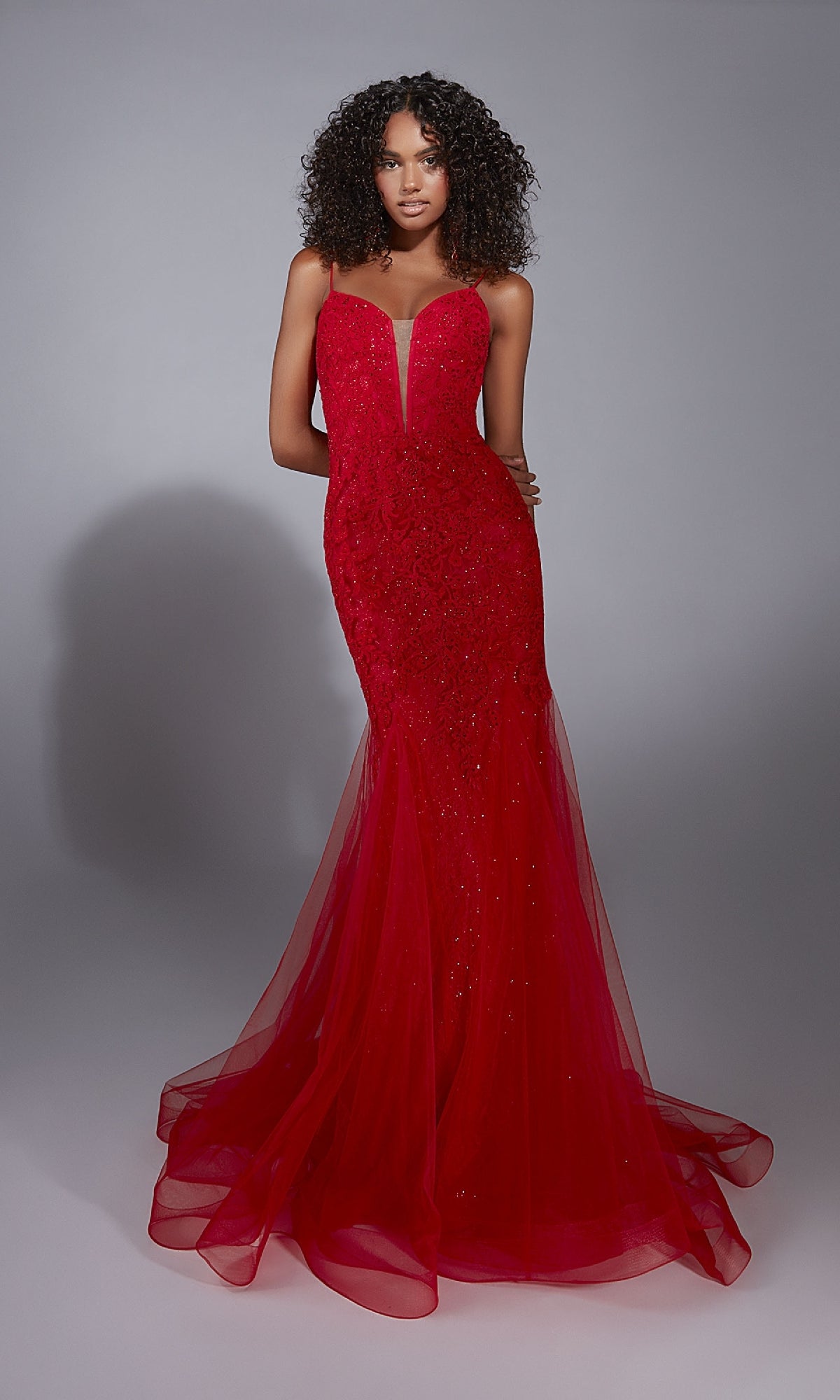 A woman stands against a gray background wearing the Aerie Couture 334, a fitted, floor-length red mermaid prom dress featuring thin straps, a deep neckline, sheer flared skirt, and an elegant open back.