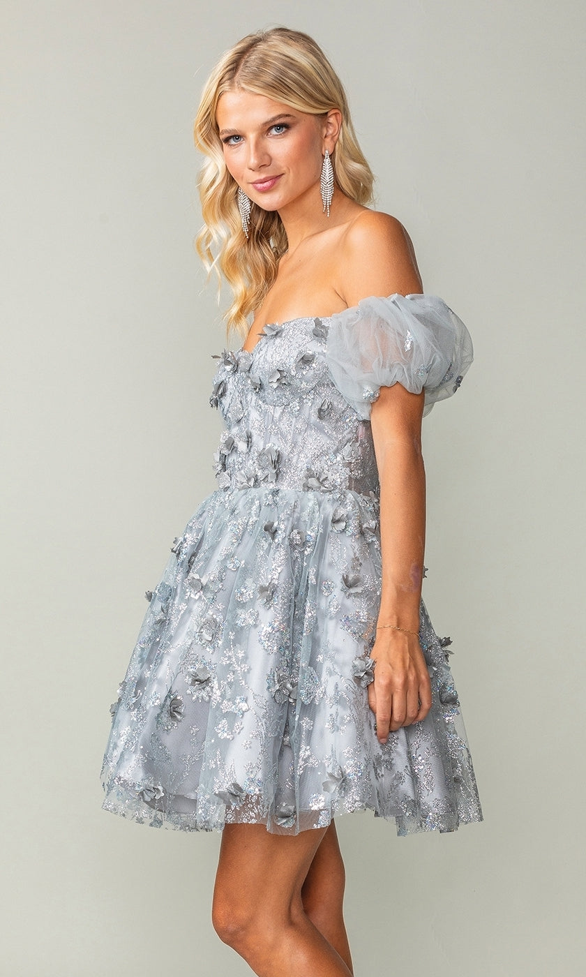 A woman wears the Short Sleeve Short Glitter Homecoming Dress 3355, a silver mini dress with puff sleeves and glitter detail, posing against a plain light background.
