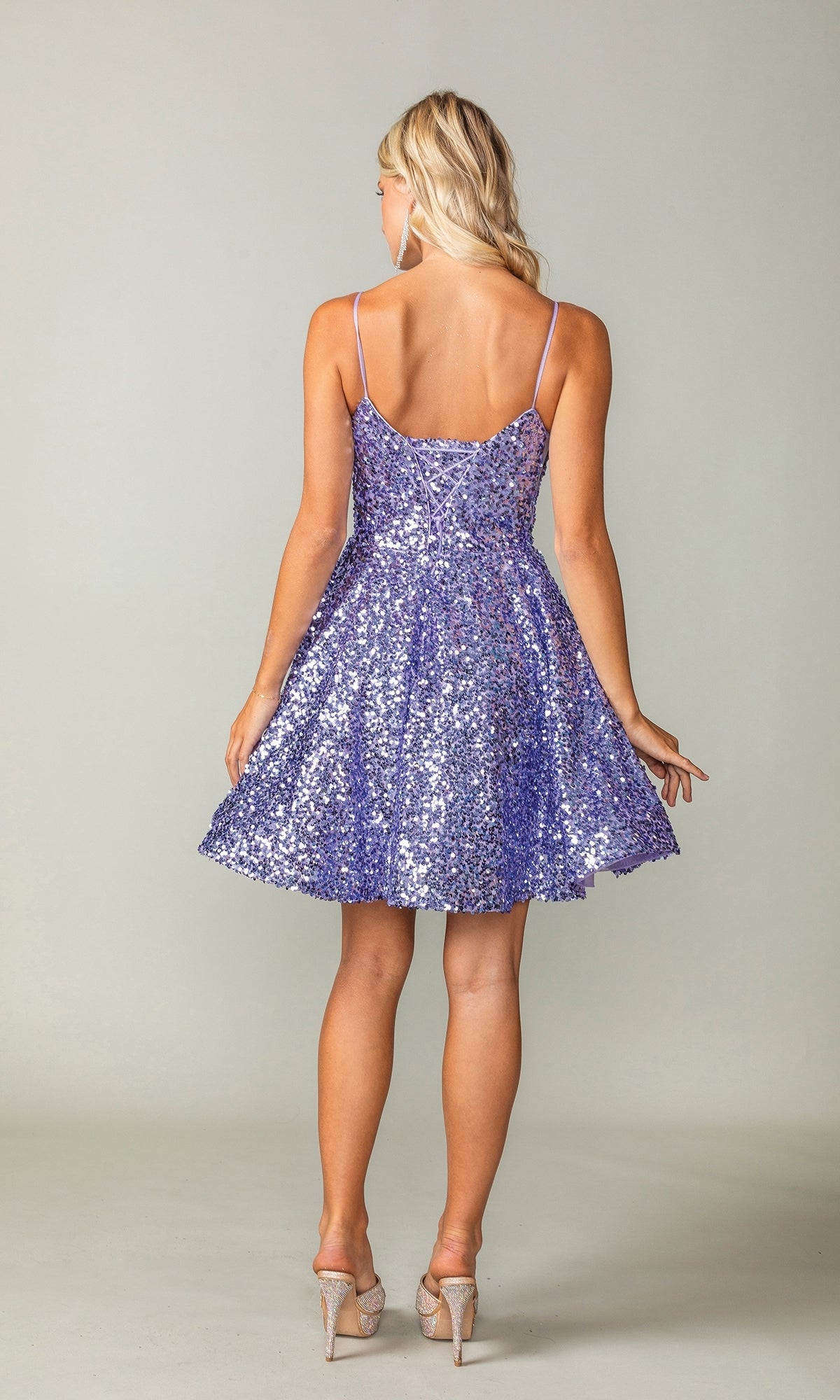 A woman faces away, wearing the Short Sequin A-Line Homecoming Dress 3356 in sparkly lavender with thin straps and flared skirt, paired with clear high-heeled shoes.