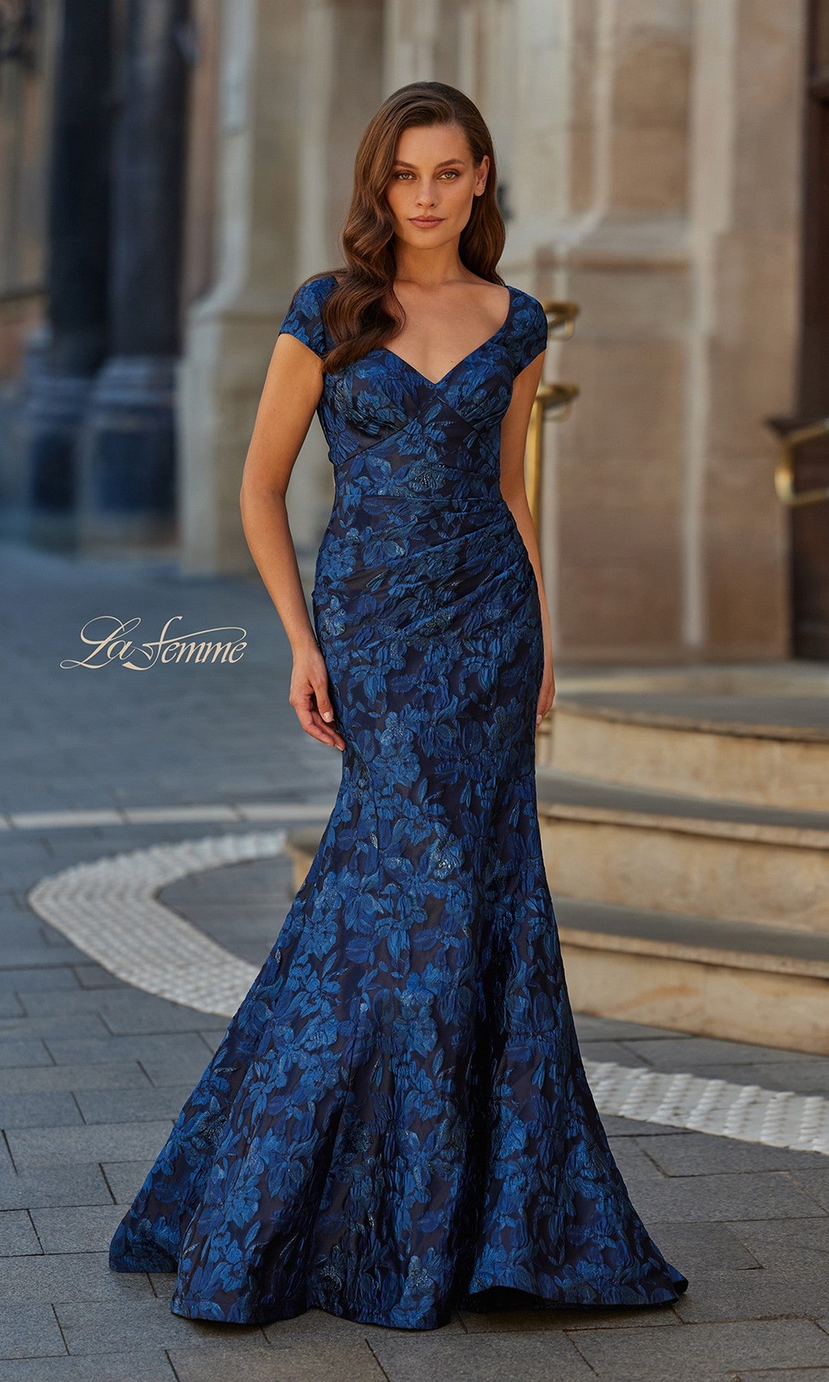 A woman stands outdoors on a stone walkway near building steps, wearing the La Femme 33578 long formal dress in navy—a fitted, floor-length mermaid gown with cap sleeves.