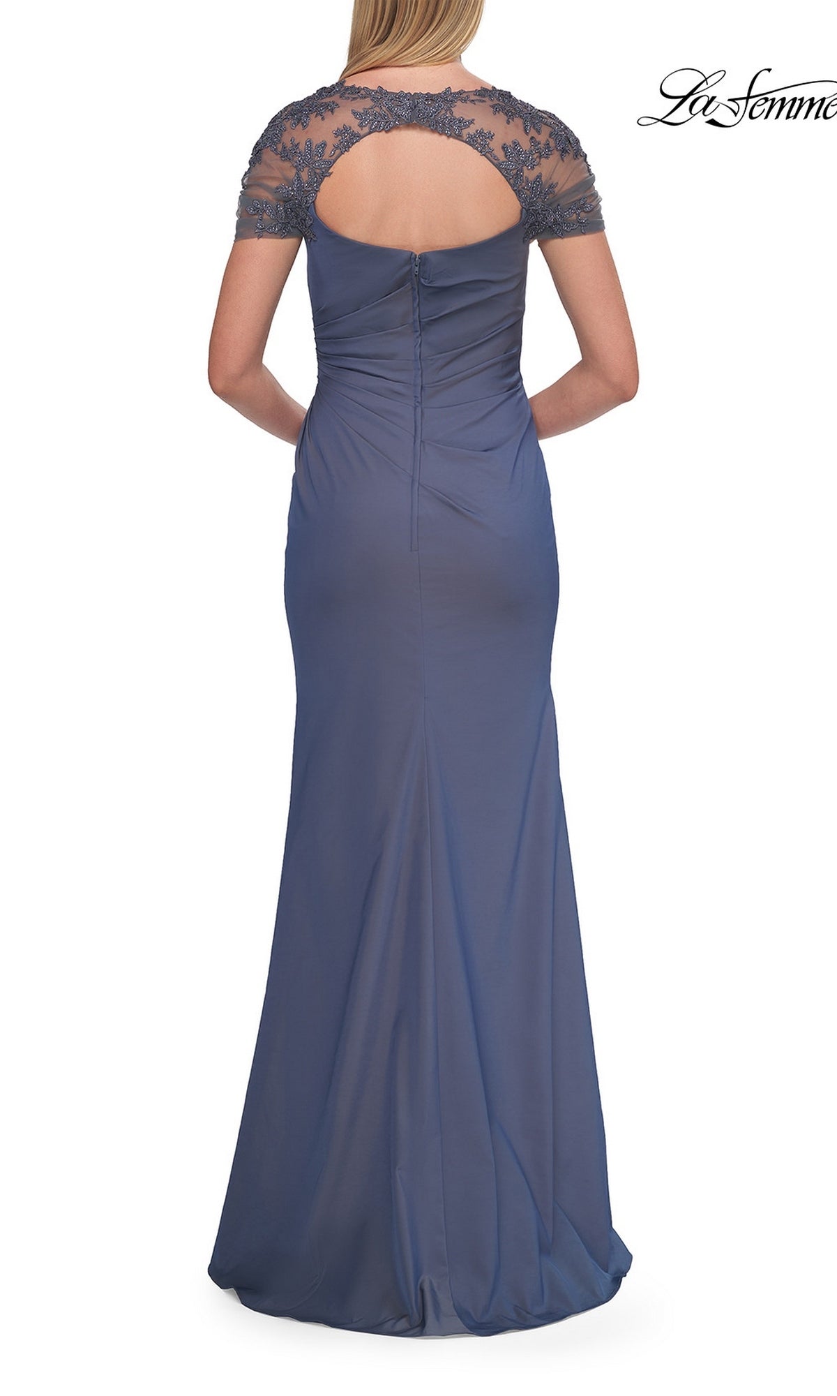 A graceful mother of the bride stands with her hands clasped behind, wearing the Back Cut-Out Long Formal Dress: La Femme 33579—a stunning blue floor-length gown featuring short lace sleeves and an elegant open-back detail.