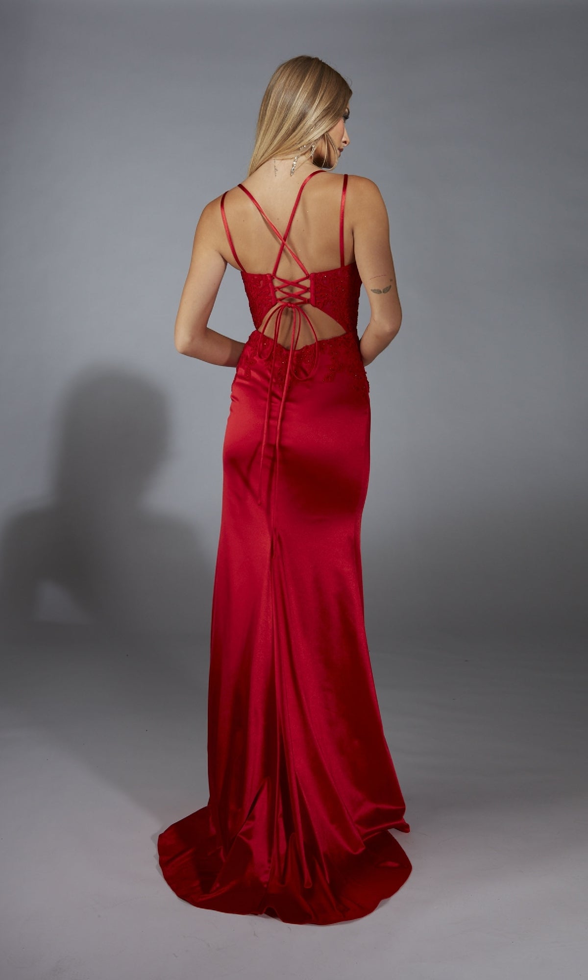A woman stands facing away, wearing the Aerie Couture 335—a long red satin prom dress featuring crisscross straps, an open back, and a graceful leg slit.
