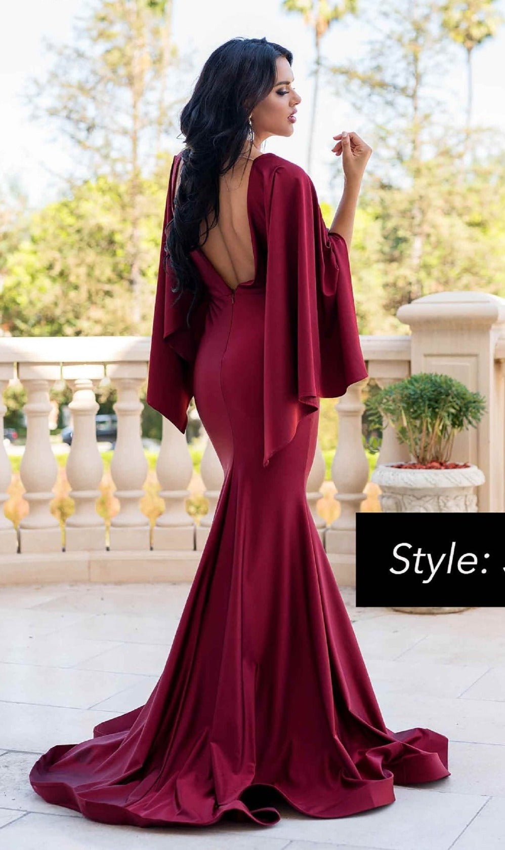A woman stands outdoors on a stone terrace wearing the Jessica Angel 336 Long Cape-Sleeve Formal Dress, a floor-length deep red gown with a low back and elegant cape-style sleeves.