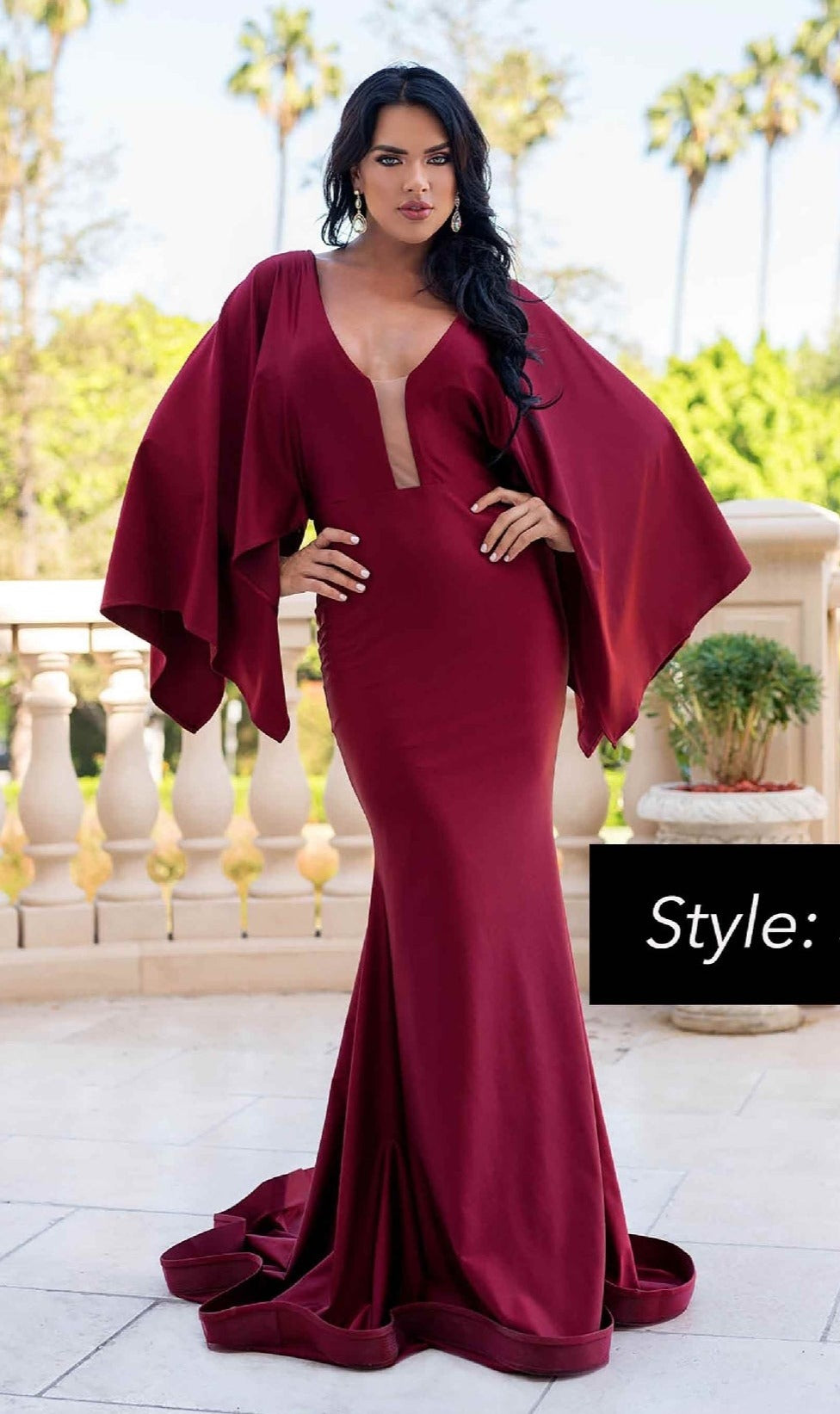 A woman in the Jessica Angel 336 Long Cape-Sleeve Formal Dress in deep red stands on a terrace, framed by lush greenery and palm trees.