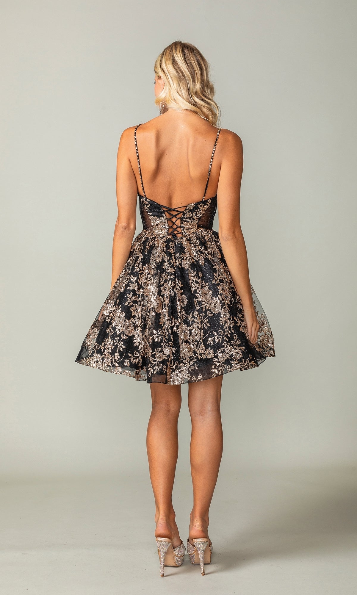 A woman faces away in the Short A-Line Glitter-Print Homecoming Dress 3360, featuring black and gold glitter print, a lace-up back, flared skirt, and high heels.