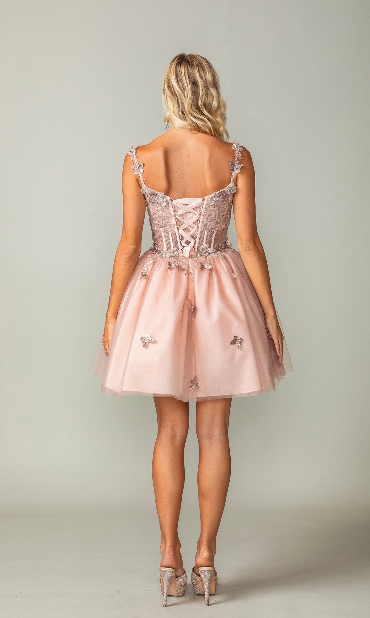 A woman faces away wearing the Rose Gold Short Butterfly Homecoming Dress 3367, featuring a lace-up back, floral appliques, and paired with high heels.