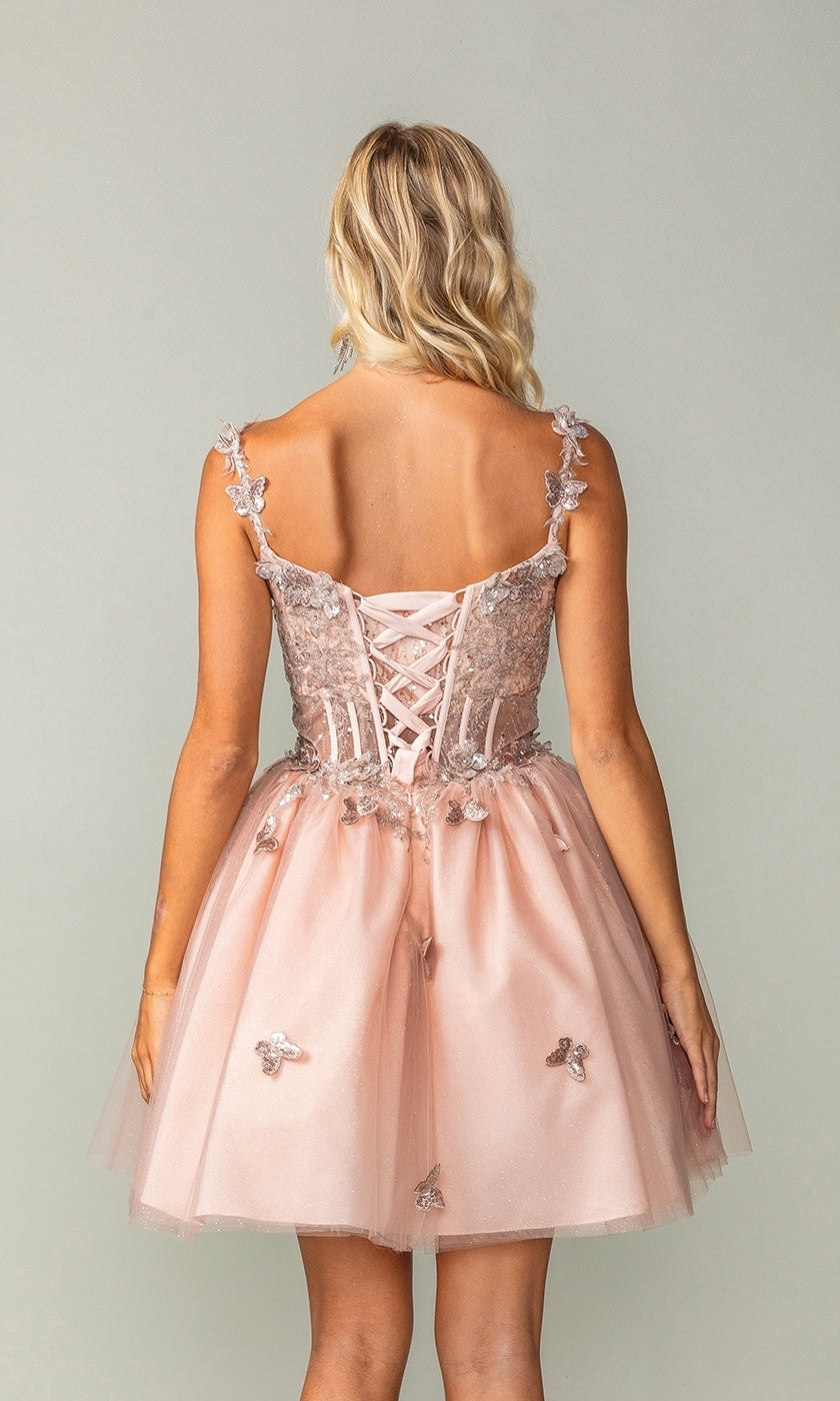 A woman with wavy blonde hair is seen from behind against a plain light background, wearing the Rose Gold Short Butterfly Homecoming Dress 3367 featuring a blush pink corset bodice and floral appliqués.