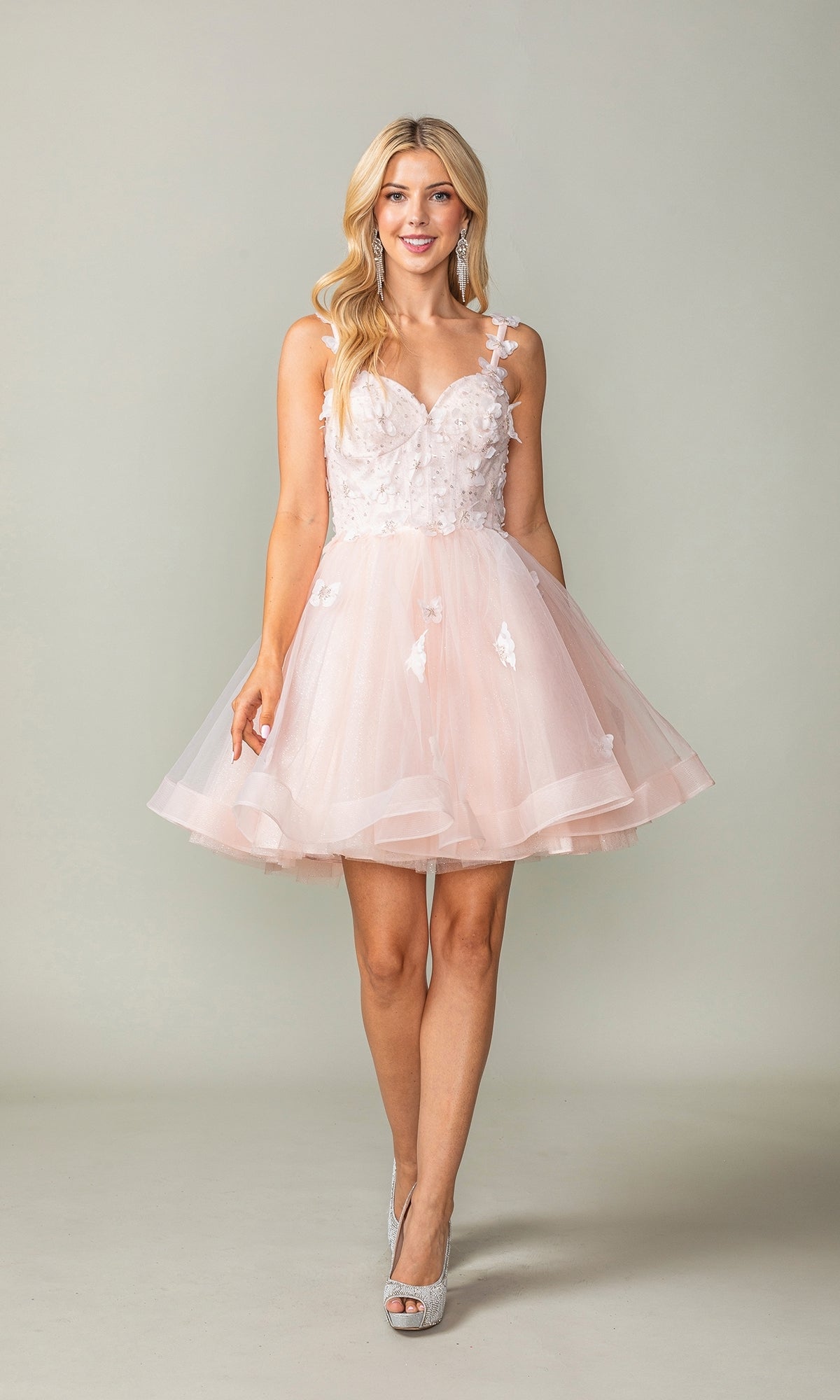 A woman in the Lace-Up Short Butterfly Homecoming Dress 3369 featuring a light pink, sleeveless design with 3-D butterflies and a tulle skirt, paired with silver heels, stands against a plain background.