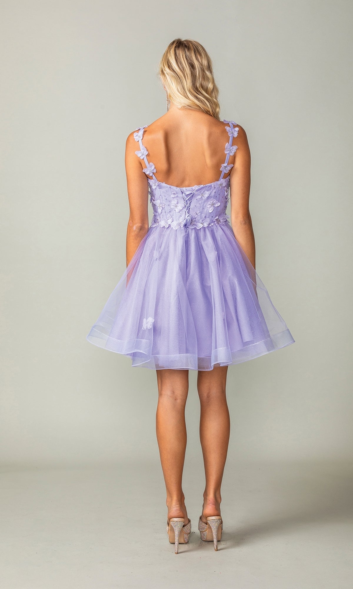 A woman faces away, wearing the Lace-Up Short Butterfly Homecoming Dress 3369 in lavender with floral accents, 3-D butterflies, sheer layers, and high heels.