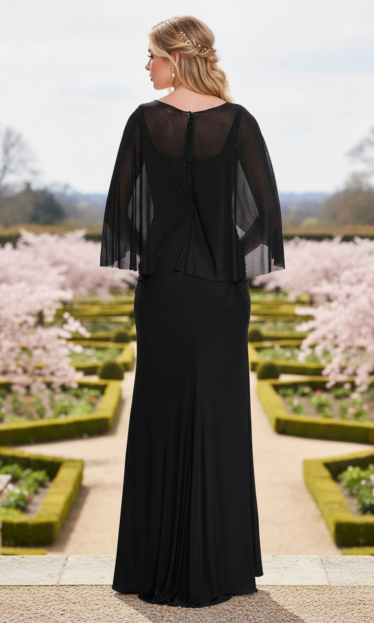 A woman in the La Femme 33718 Long Prom Dress with sheer sleeves stands outdoors on a garden path, facing away from the camera toward blooming trees and manicured hedges.