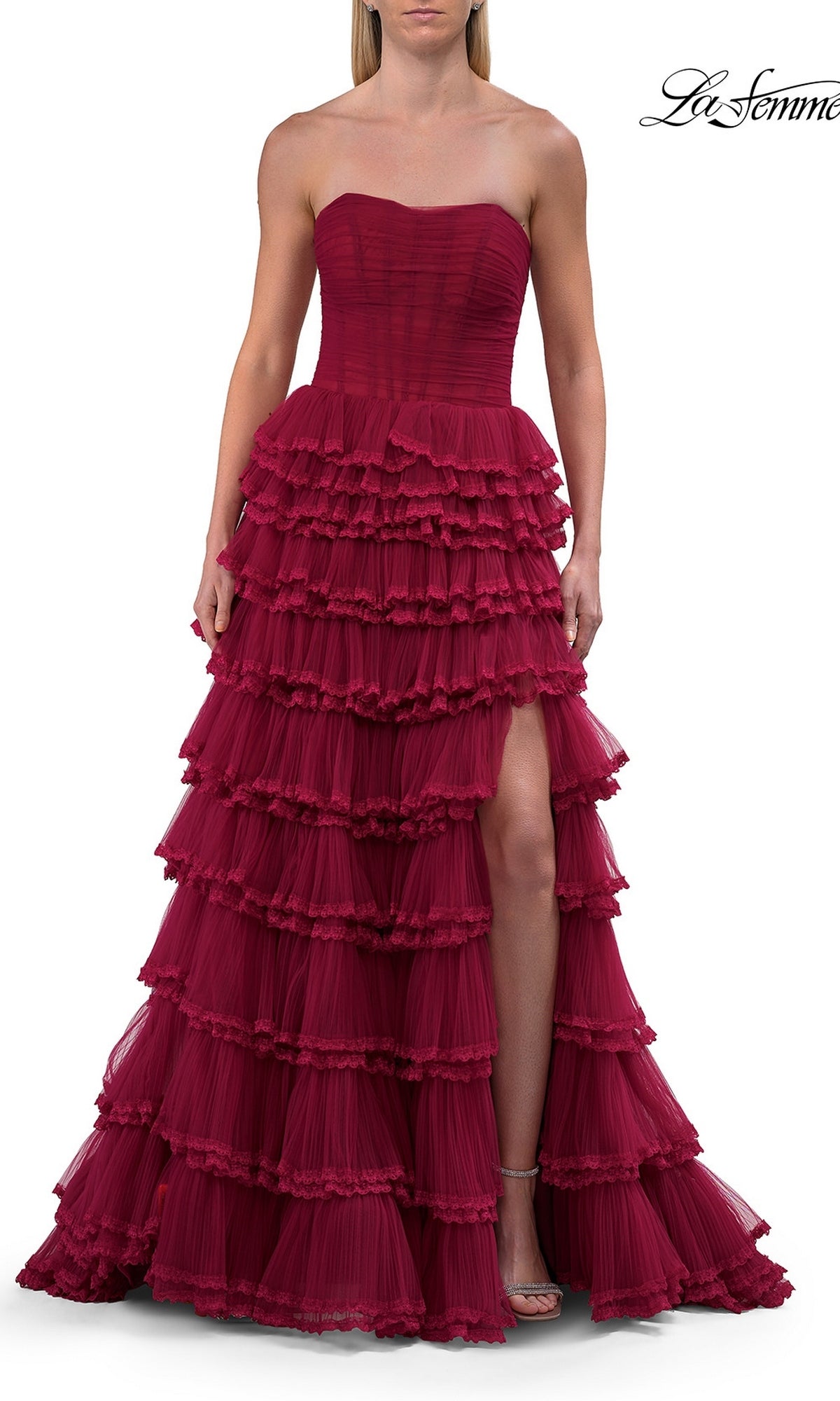 A woman wearing the La Femme 33727 long prom dress in raspberry, a strapless tulle ball gown with ruffled tiered layers, a corset bodice, and a high slit, standing against a white background.