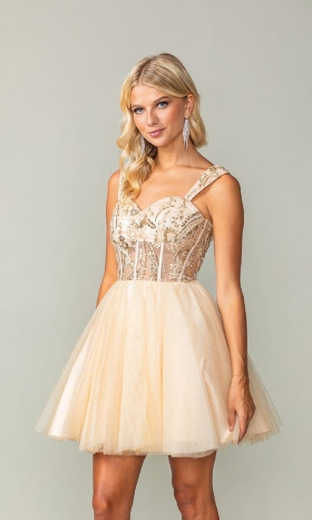 A woman in a champagne Lace-Up Open-Back Hoco Dress (Dancing Queen 3375), featuring sleeveless tulle and sequin embellishments, stands against a plain background.