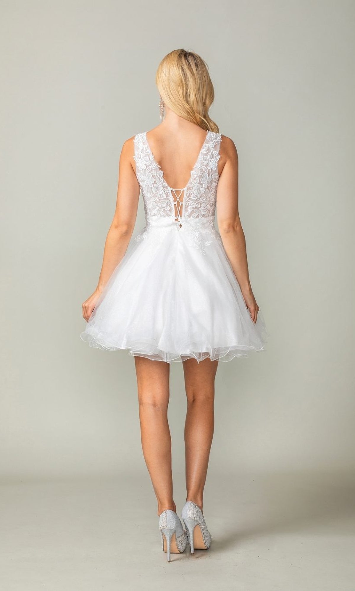 A woman models the Tulle Babydoll Homecoming Dress: Dancing Queen 3377, a short, sleeveless white dress with lace details and a lace-up low back, standing in high heels facing away from the camera.