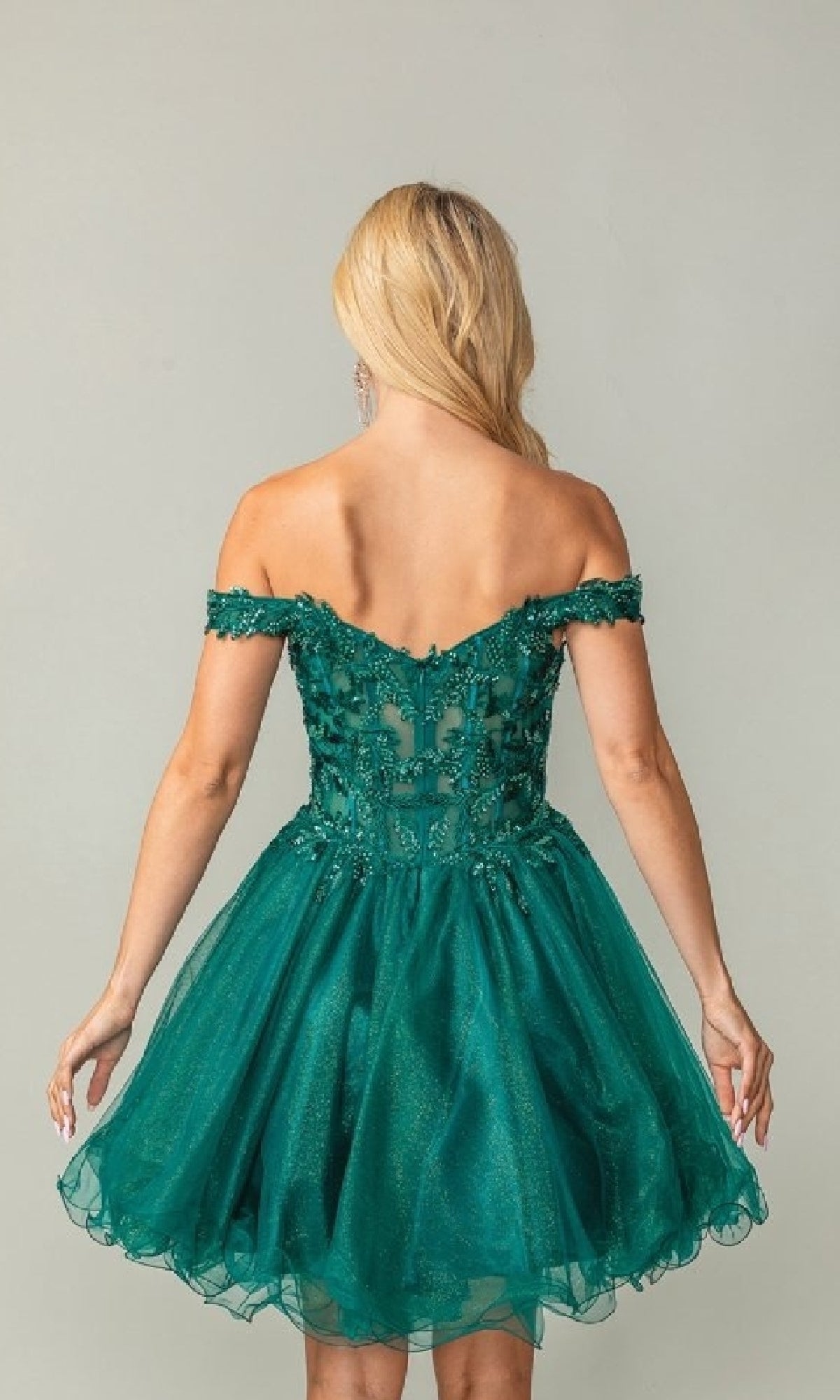 Woman showing the back of a dark green short off-shoulder dress with lace details on a plain background