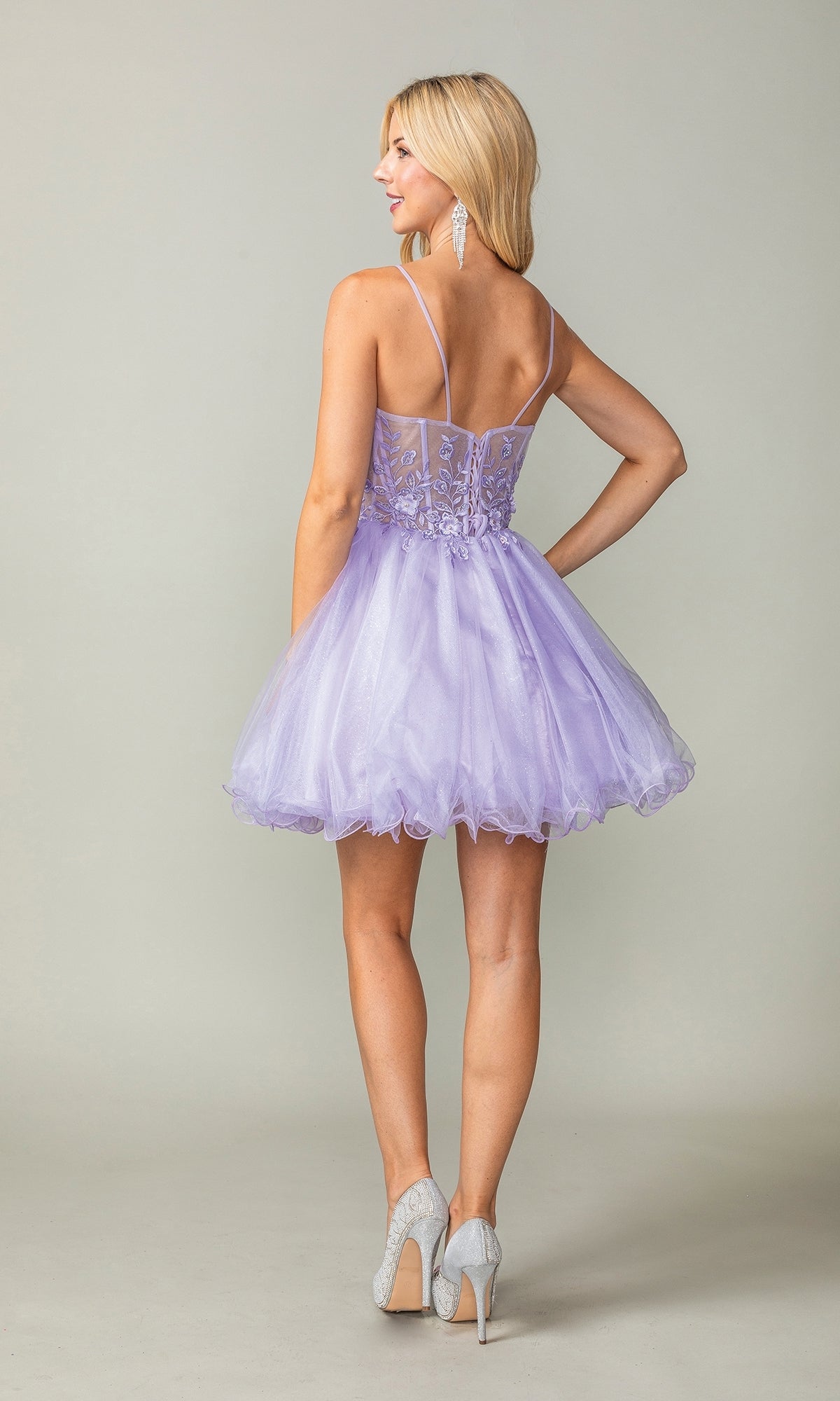 A woman stands facing away, wearing the Sheer-Waist Short Babydoll Homecoming Dress 3382—a light purple dress with a sheer lace bodice, spaghetti straps, and tulle skirt—paired with silver high heels and long earrings.