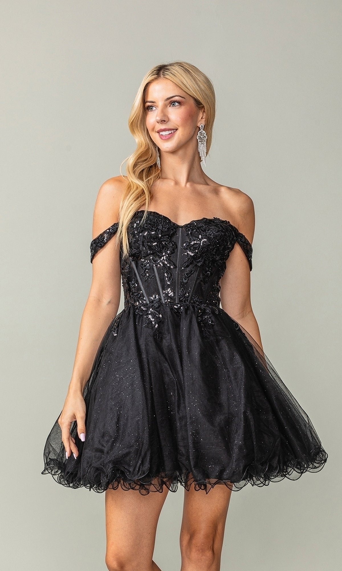 A woman stands against a plain background wearing the Short Corset Homecoming Dress: Dancing Queen 3383—a black, off-the-shoulder, short tulle dress featuring floral embellishments and a sparkly sheer corset bodice.