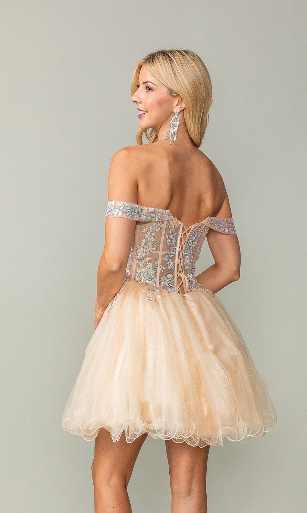 A woman stands against a plain background wearing the Dancing Queen 3383 short corset homecoming dress featuring an off-shoulder neckline, lace-up back, floral embroidery, and a tulle skirt.