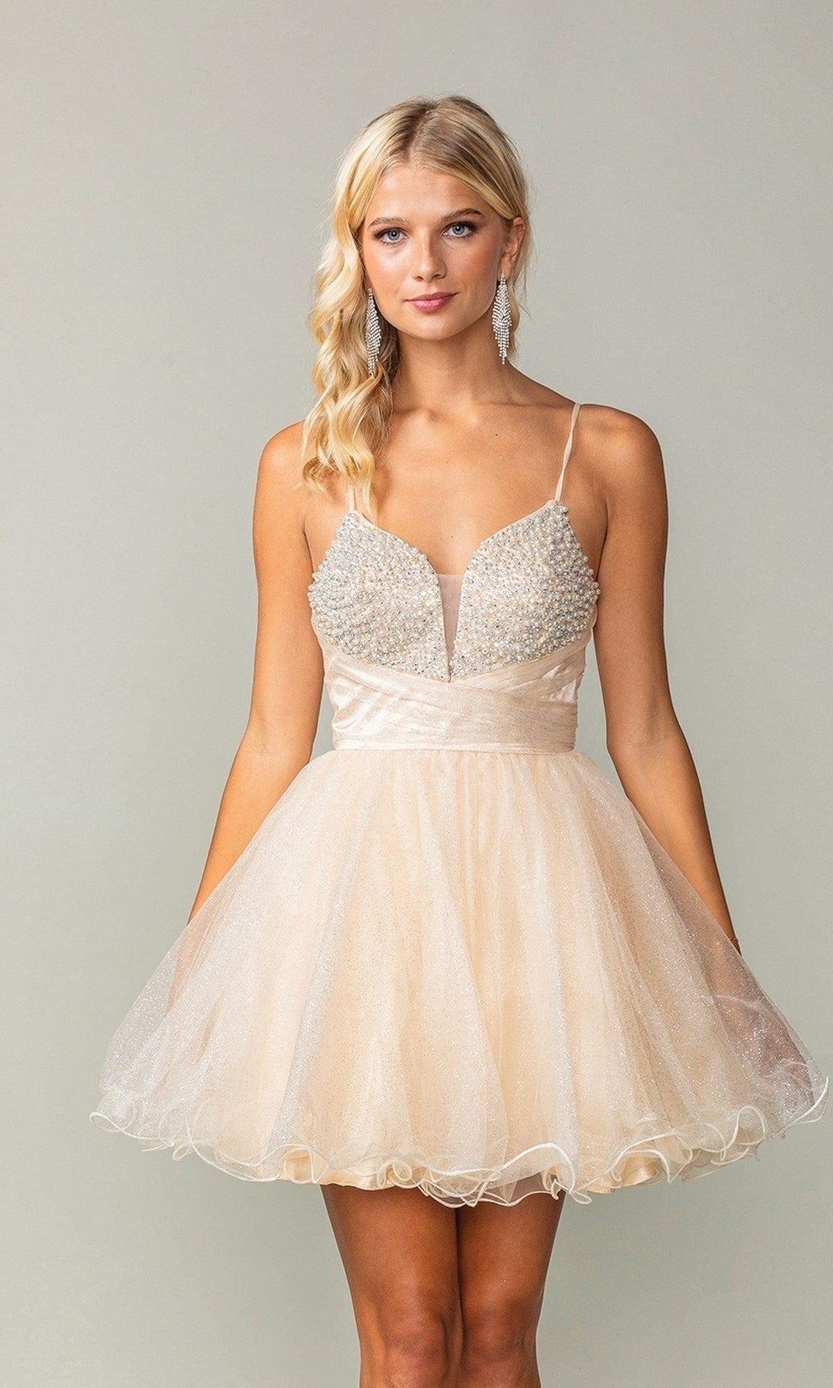 A woman models the Lace-Up Short Babydoll Homecoming Dress 3385, an affordable light beige cocktail dress with a fitted jeweled bodice and flared tulle skirt, standing against a plain background.
