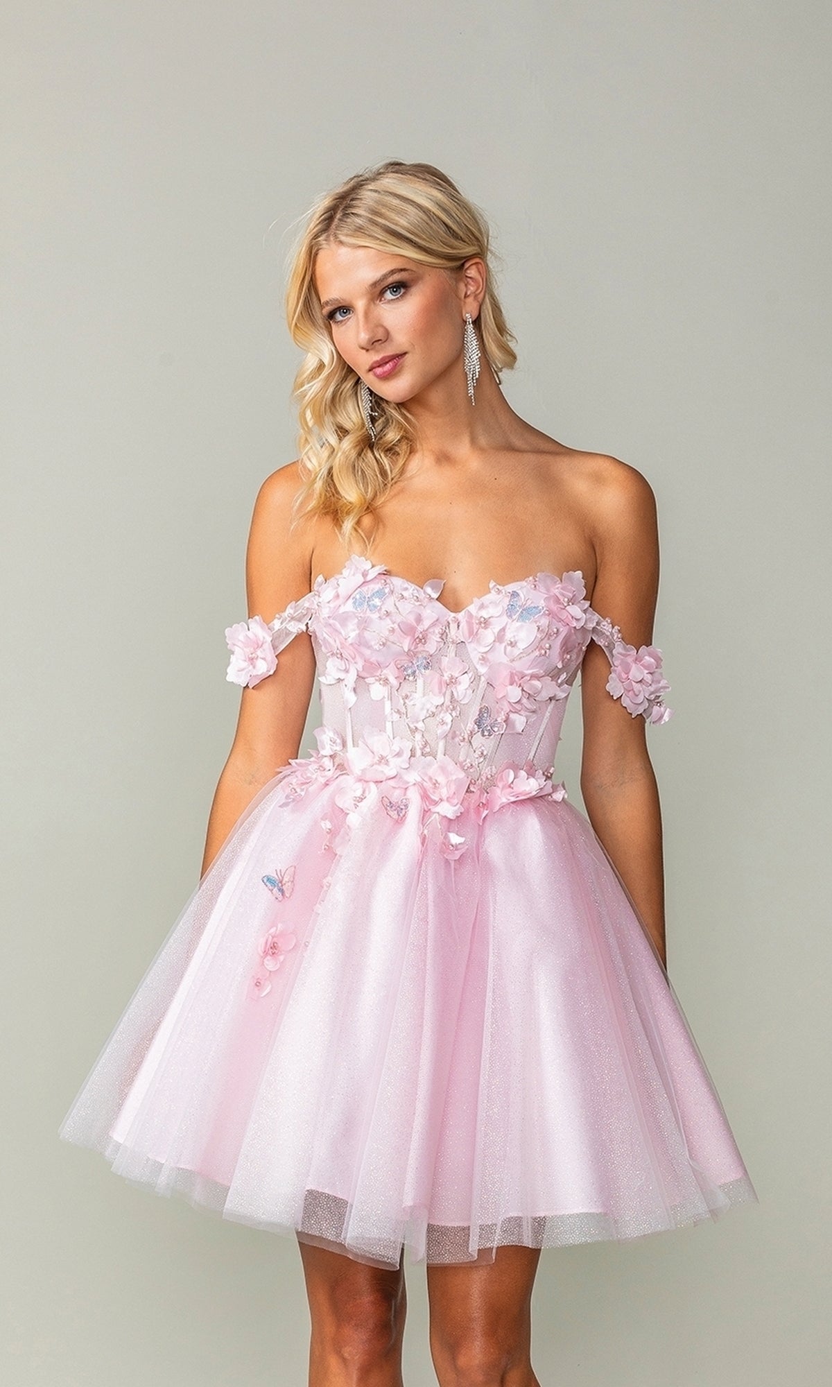 A woman models the Off-Shoulder Short Babydoll Homecoming Dress 3388—a strapless, pink dress with floral appliques and a tulle skirt—while standing against a neutral background.