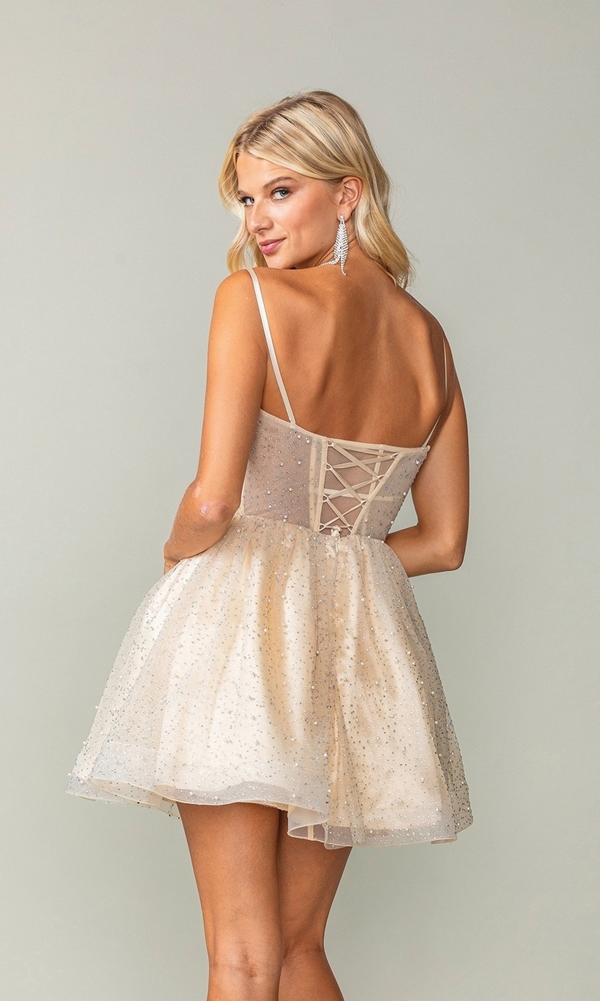 A woman wearing the Short Glitter Fit-and-Flare Homecoming Dress 3389, featuring a sparkly light hue and lace-up back, stands facing away from the camera, looking over her shoulder.
