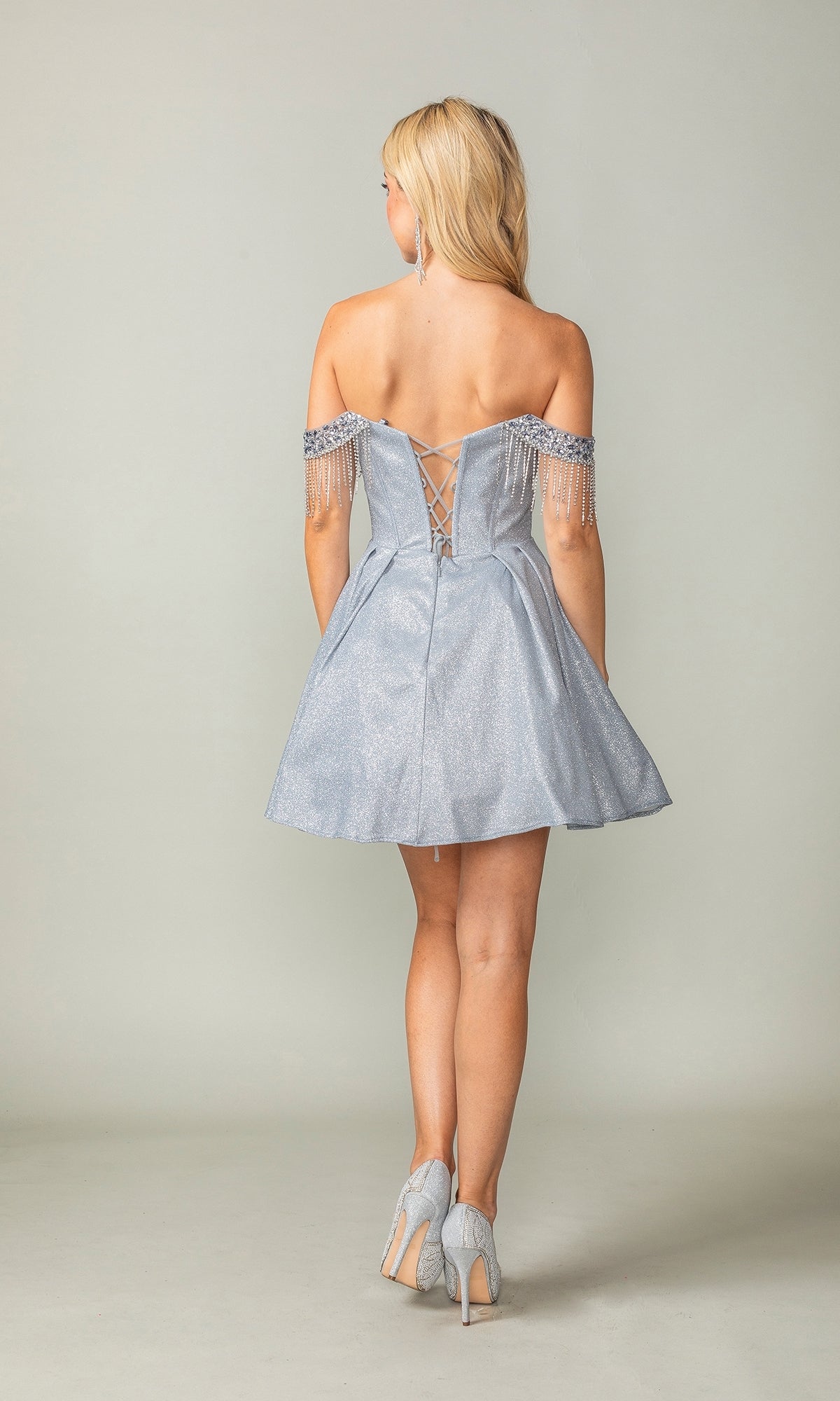 A woman faces away from the camera, wearing the Fringed Off-Shoulder Short Homecoming Dress 3392, which features an off-the-shoulder style, lace-up corset bodice, beaded sleeves, and silver heels.