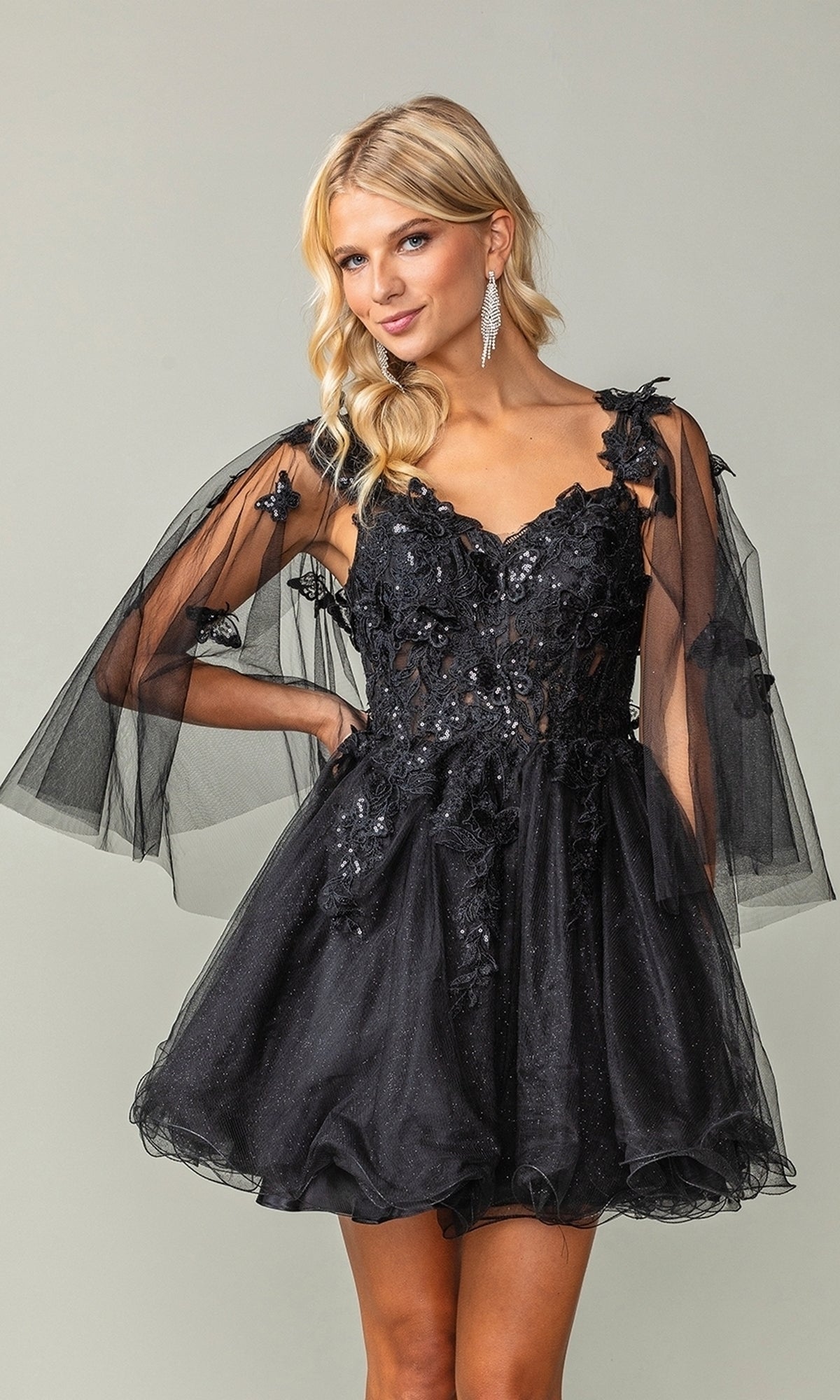 A woman models the Butterfly-Cape Short A-Line Homecoming Dress 3393, featuring sequin appliques and sheer, butterfly-inspired capelet sleeves, standing against a plain background.