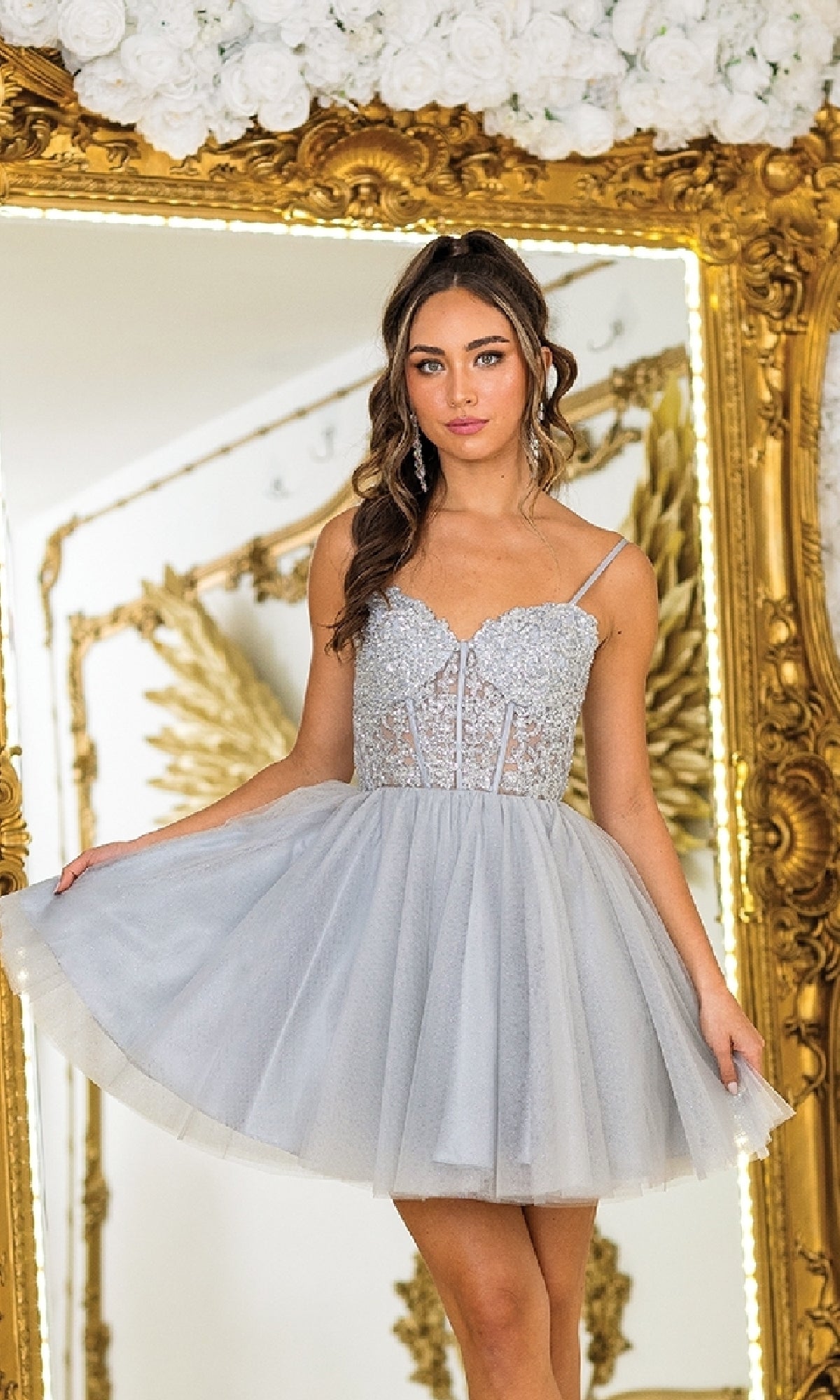 A woman wearing the Short Corset Homecoming Dress: Dancing Queen 3399, featuring a silver tulle beaded corset bodice, poses before an ornate gold mirror decorated with white flowers.