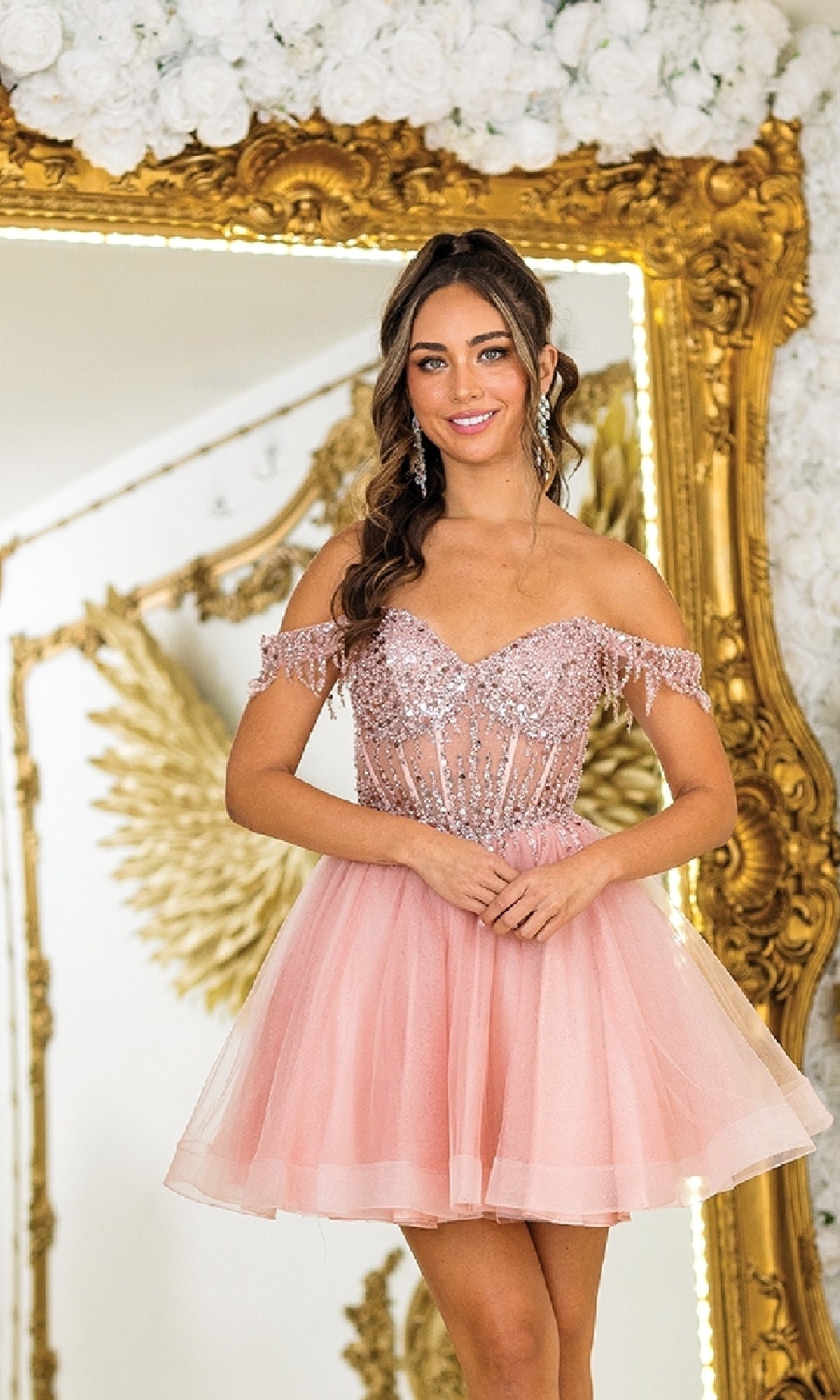 A woman in the Short Glitter Babydoll Prom Dress: Dancing Queen 3405 stands before an ornate gold-framed mirror, with gold wing decor glowing in the background.