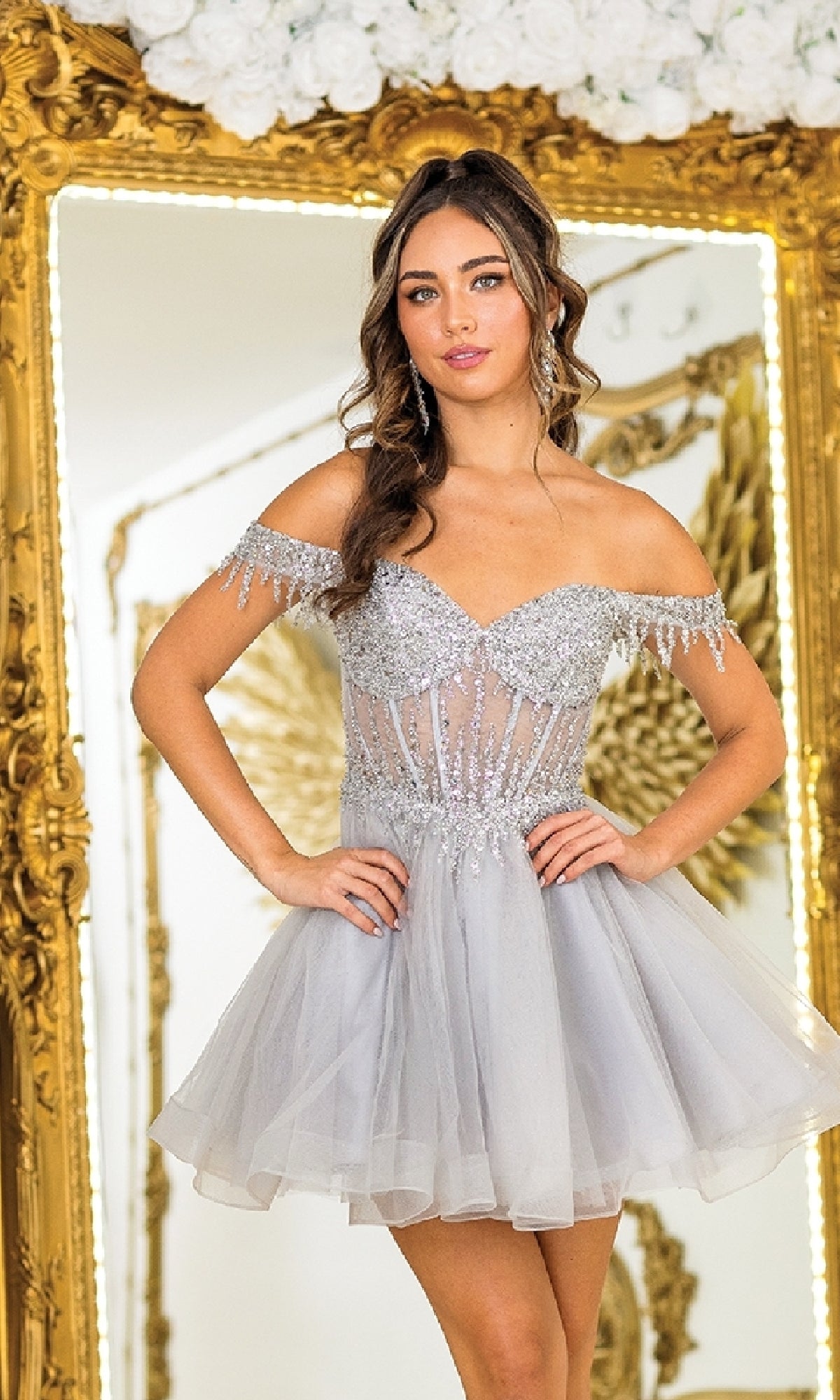 A woman in the Short Glitter Babydoll Prom Dress: Dancing Queen 3405, featuring an off-the-shoulder neckline, tulle skirt, and sparkling details, stands before an ornate gold-framed mirror.